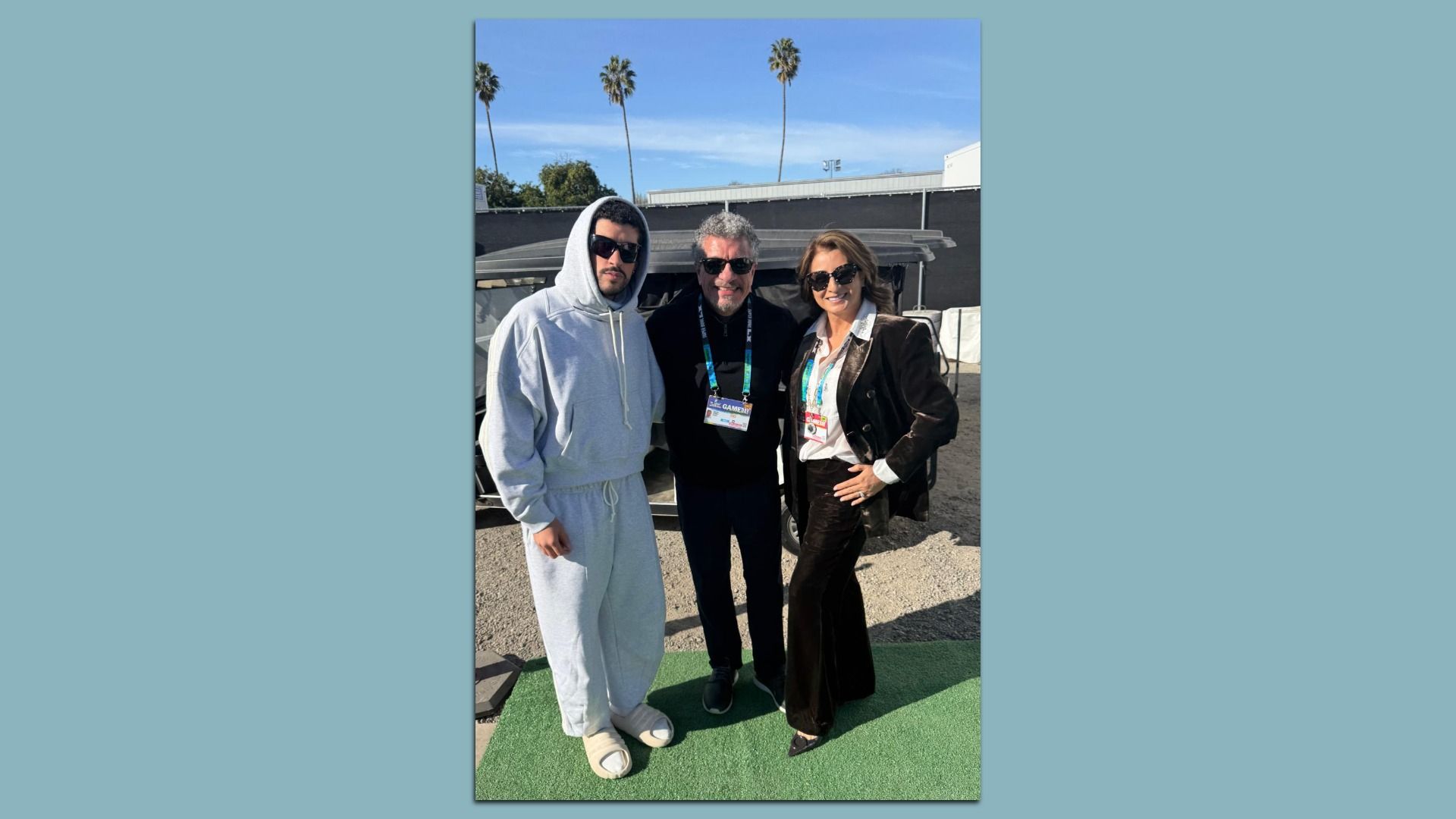 Three people wearing sunglasses stand together outdoors on green carpet. Music star Bad Bunny wears gray hoodie and pants with sandals, maestro Giancarlo Guerrero in black outfit, and another in brown suit with white shirt. Palm trees in background.