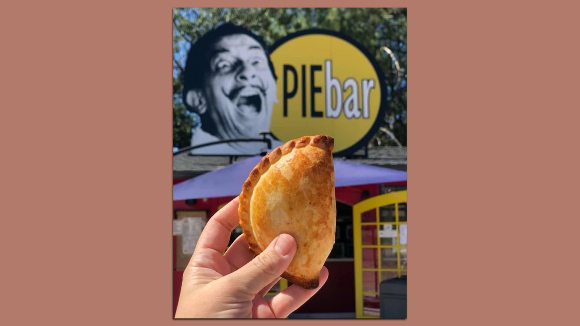 Hand holding an empanada in front of a restaurant with a sign that reads "PIEbar" next to an image of Salvador Dali's face. 