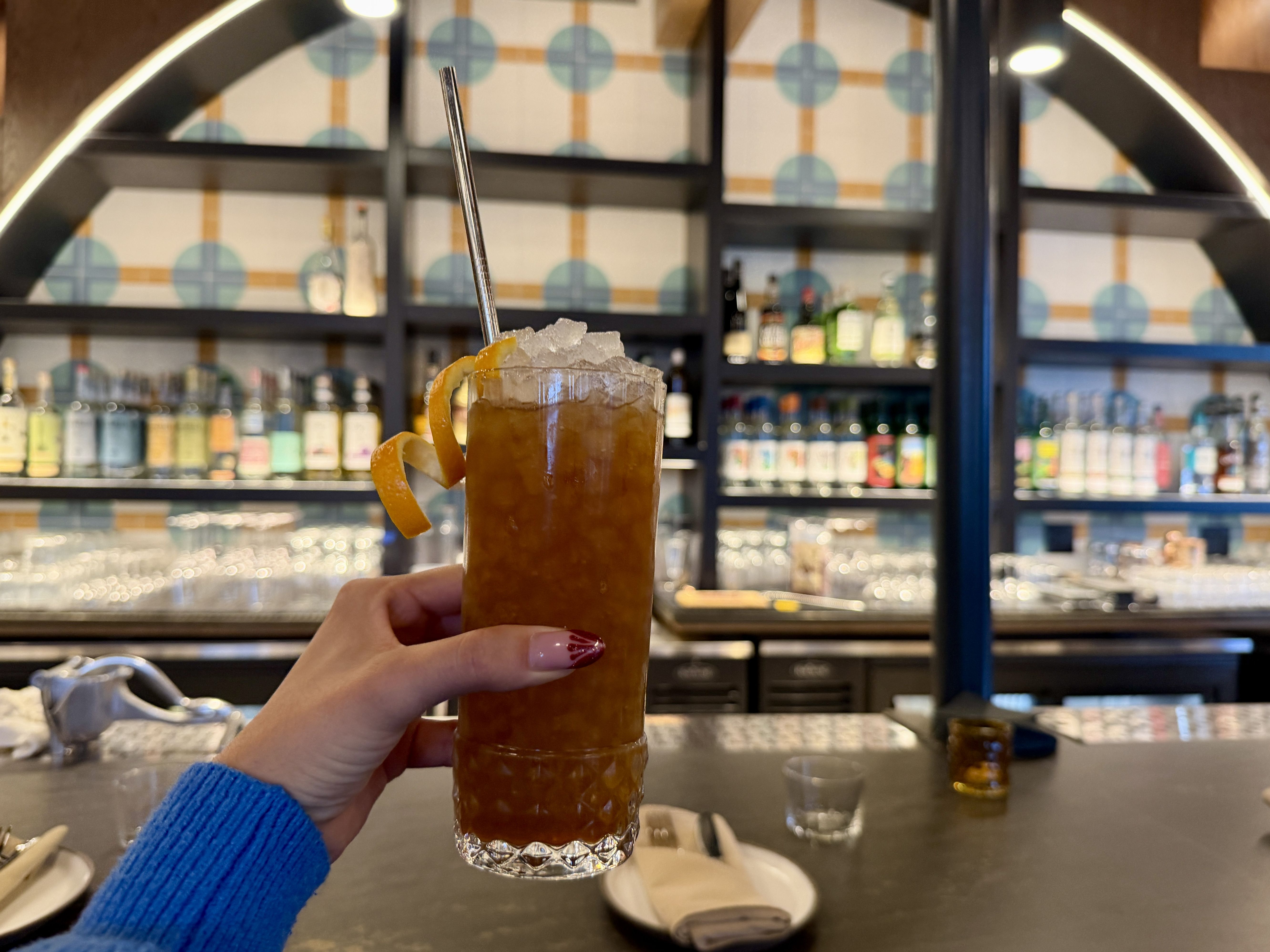 A hand with burgundy nail polish and a blue sleeve holds a tall glass of iced brown cocktail with an orange twist garnish at a bar with bottles and glasses in the background.