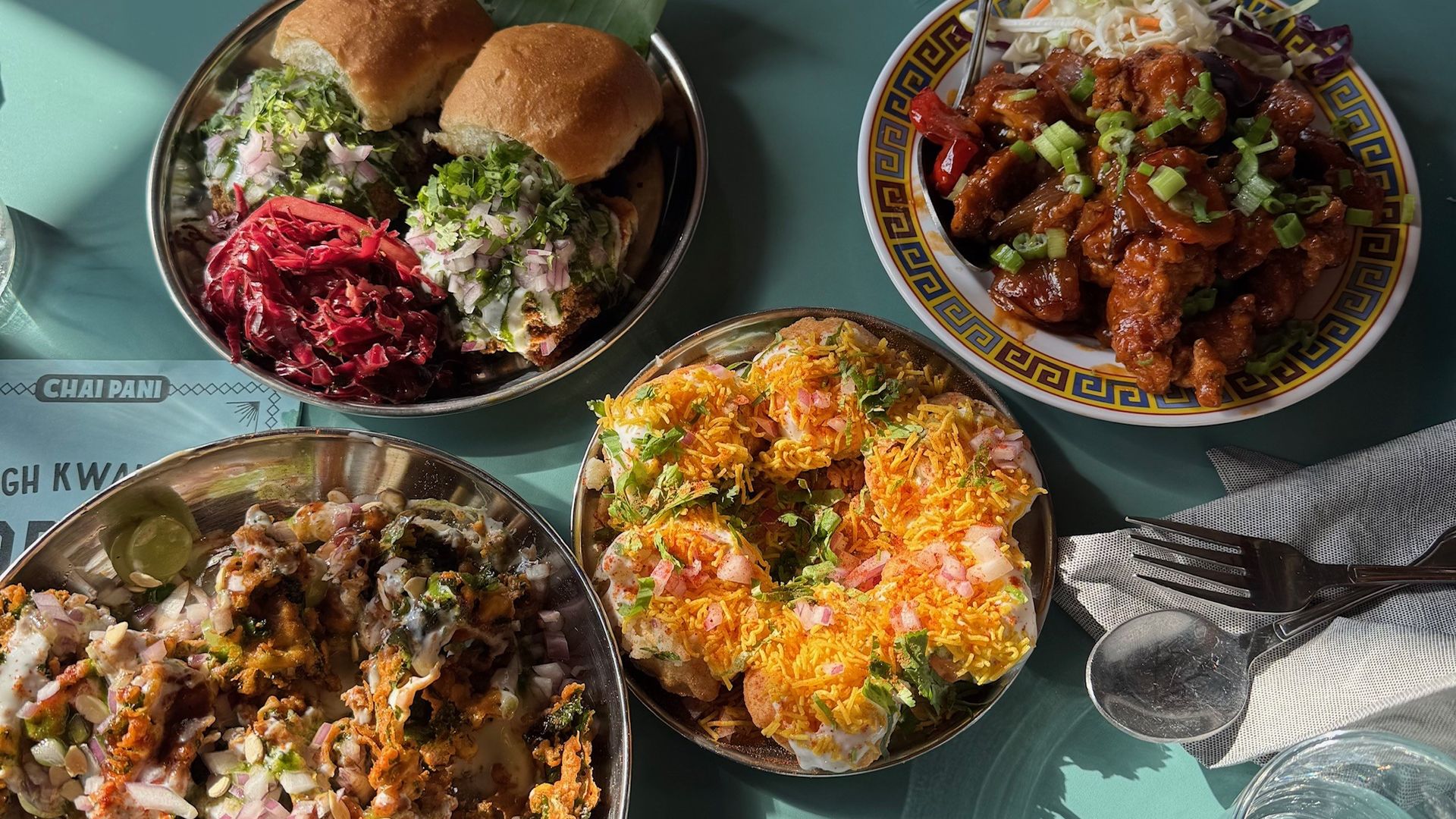 Top-down view of a teal table featuring assorted dishes: two mini burgers with greens, a plate of saucy fried chicken bites with scallions, a bowl of colorful chaat with yogurt and onions, and a cheesy, topped dish.