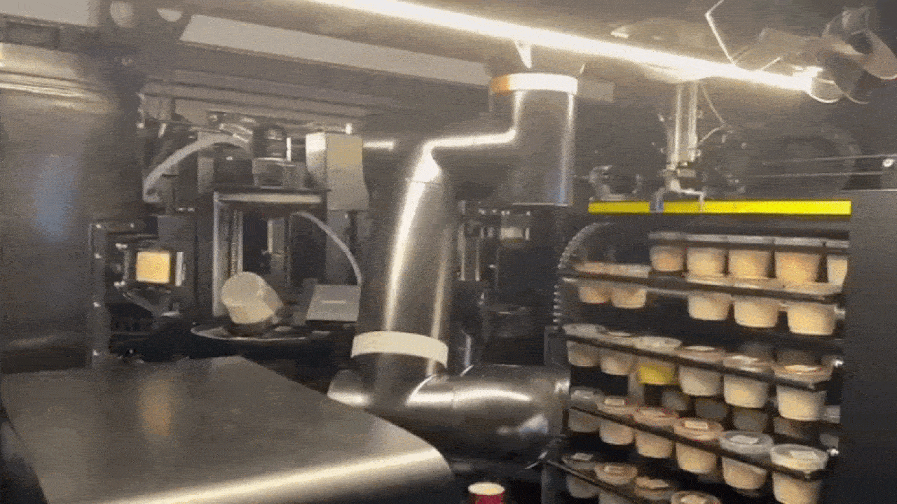 Robot arm in an industrial setting picks ice cream cups from conveyor and stacks them on shelves. The environment is metallic with bright lighting above.