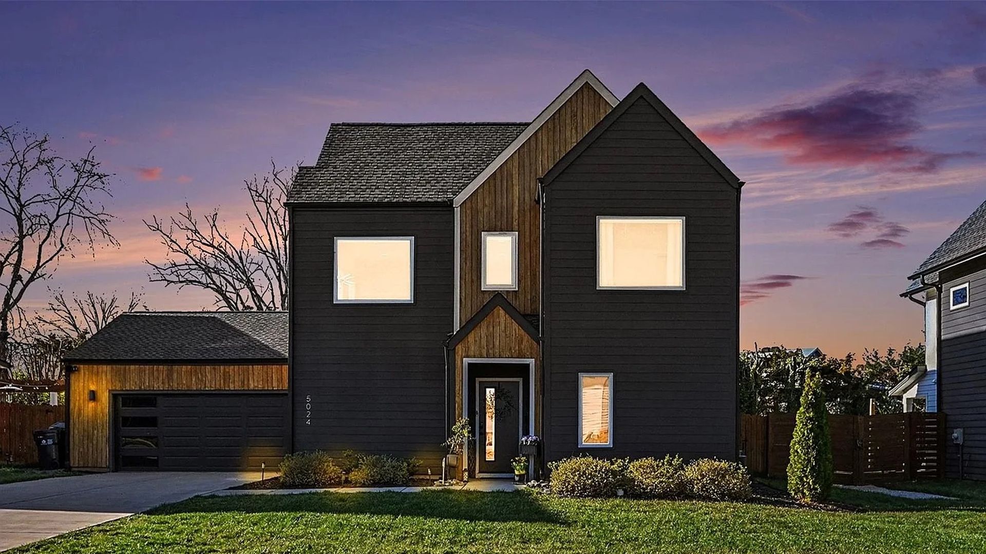 Two-story dark siding house with a wooden central gable accent, bright front windows, and a small porch. A two-car garage on the left; green lawn beneath a purple sunset sky.