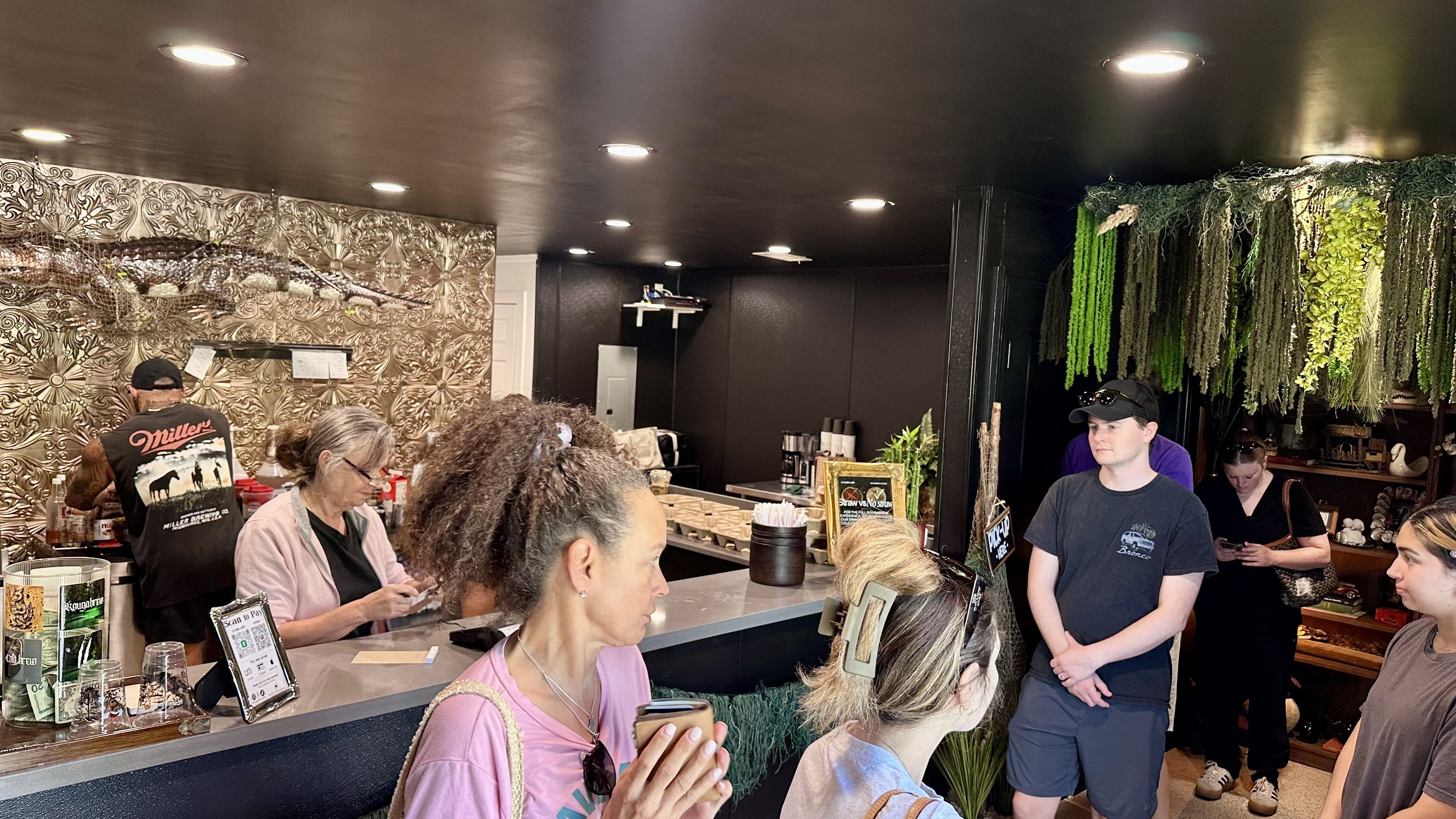 A busy cafe interior with a dark ceiling and recessed lights. On the left is an ornate silver embossed wall; on the right hangs green vines and plants, while customers line up at the counter.