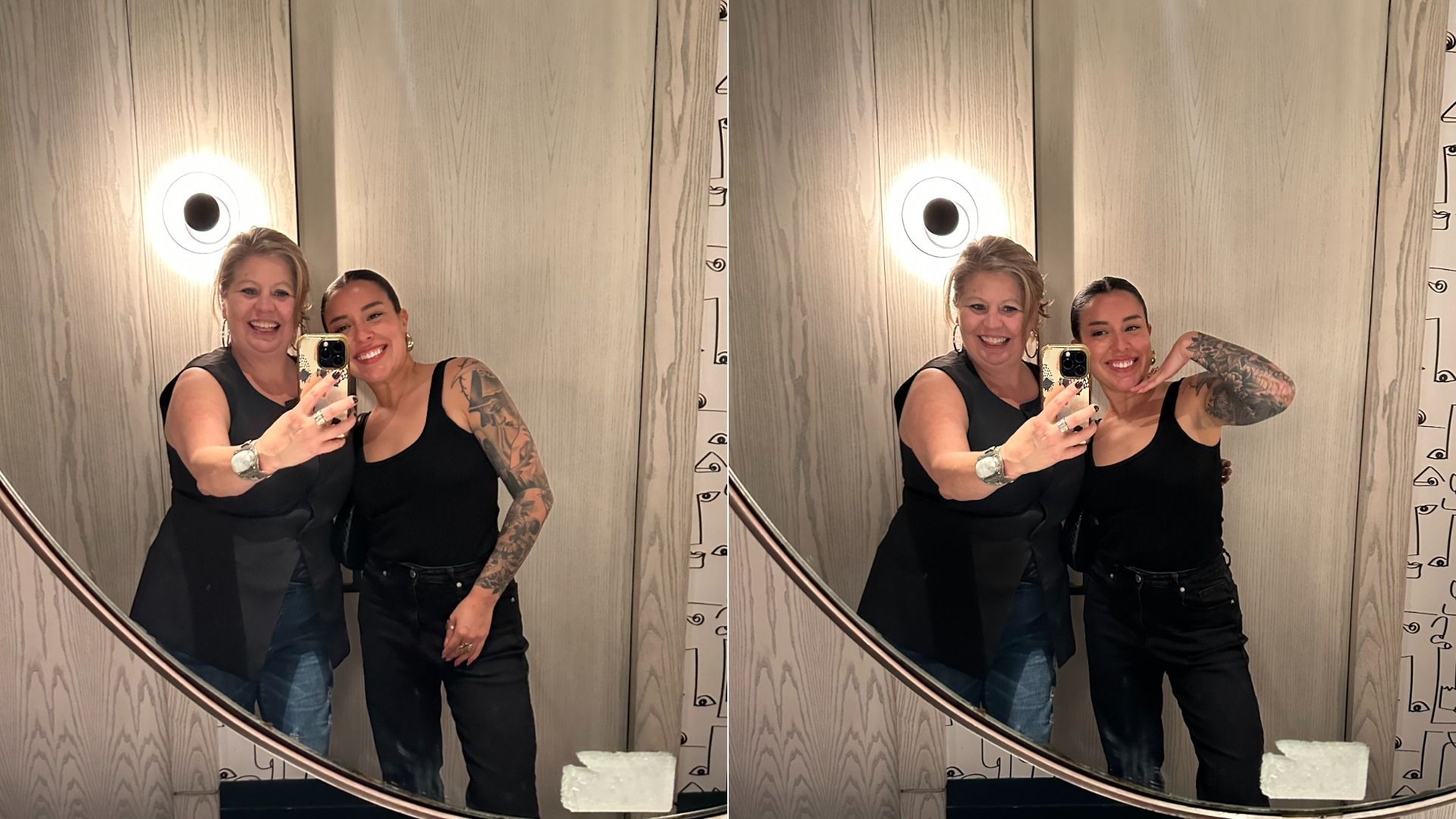 Two women smiling and taking mirror selfies in a bathroom with wood-paneled walls and a round light. One has blonde hair, black sleeveless top, jeans; the other has tattoos, black tank, and pants.