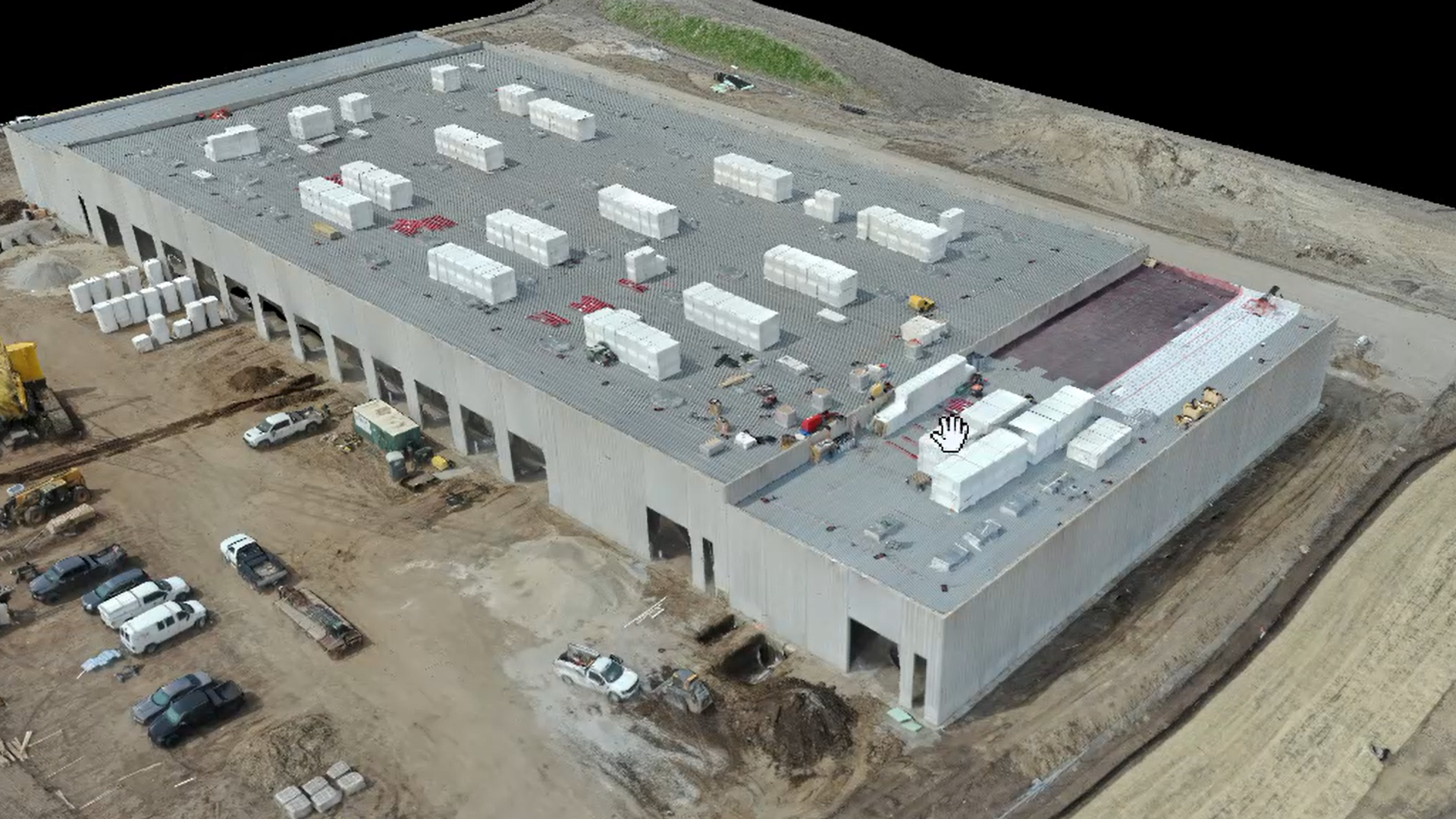 Aerial view of a large construction site with a rectangular building under construction, materials and vehicles around, and dirt roads surrounding the site.