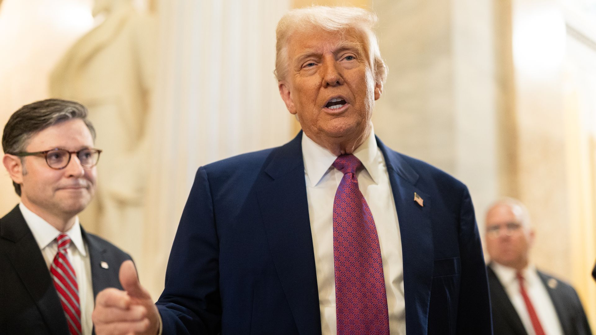 Donald Trump, wearing a blue suit and standing next to Mike Johnson in a well-lit white marble hallway.