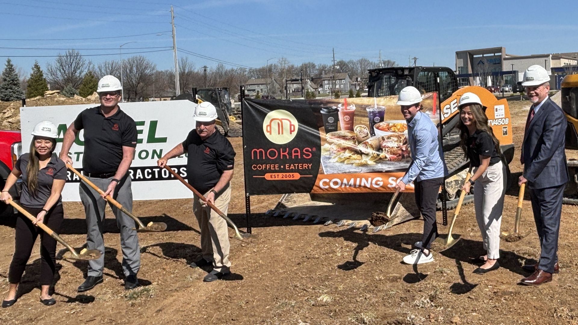 Six people in white hard hats stand with shovels at a construction site. A Mokas Coffee Eatery banner and a Coming Soon sign appear behind them, with heavy equipment in the background.