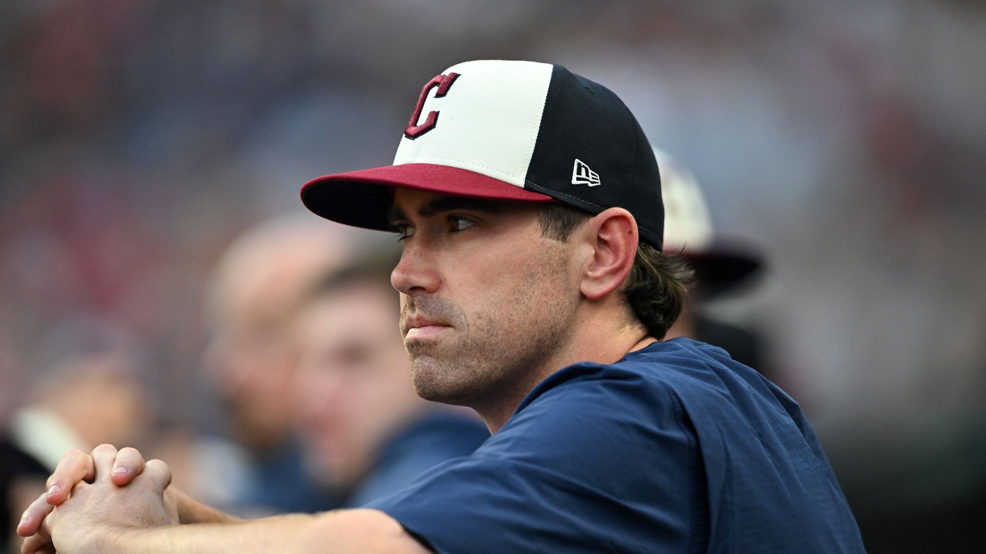 Man in a black, white, and red baseball cap with a 'C' on it, wearing a navy blue shirt, resting arms folded on a surface, looking intently to the side with blurred background.