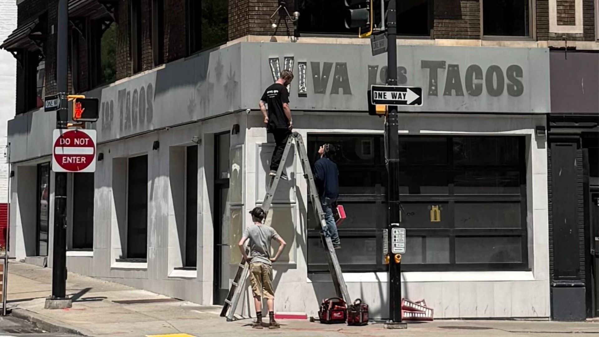 Urban street corner with a gray storefront reading TACOS in faded letters. Two workers on a ladder repair the sign while a man watches from below. Do Not Enter and One Way signs with a traffic light.