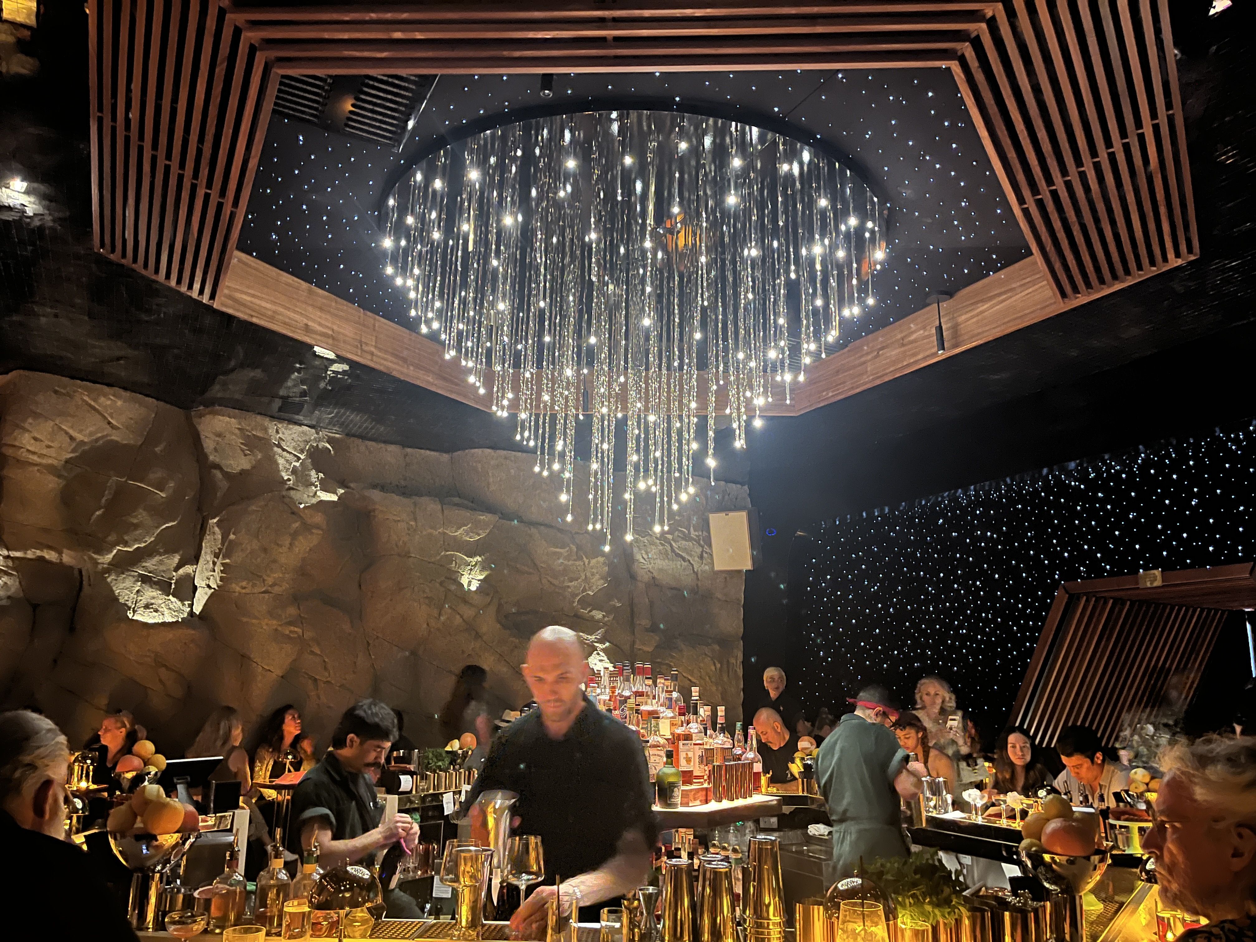 Interior of a dimly lit bar with a glowing chandelier of hanging lights, rock wall, wooden ceiling accents, bartenders preparing drinks, and customers seated at the bar under starry ceiling lights.