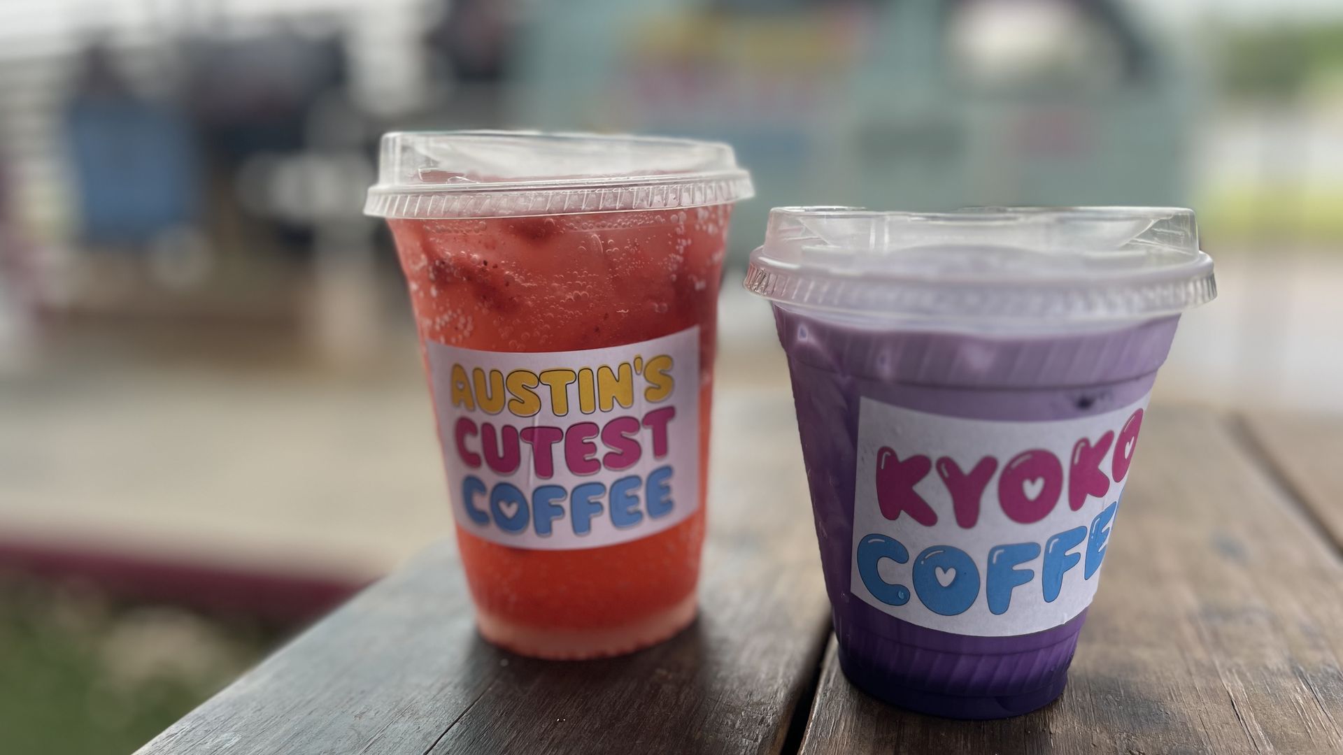 Two iced coffee cups with clear lids sit on a weathered wooden table; a red cup labeled AUSTIN'S CUTEST COFFEE and a purple cup beside it, both with condensation, blurred outdoor background.