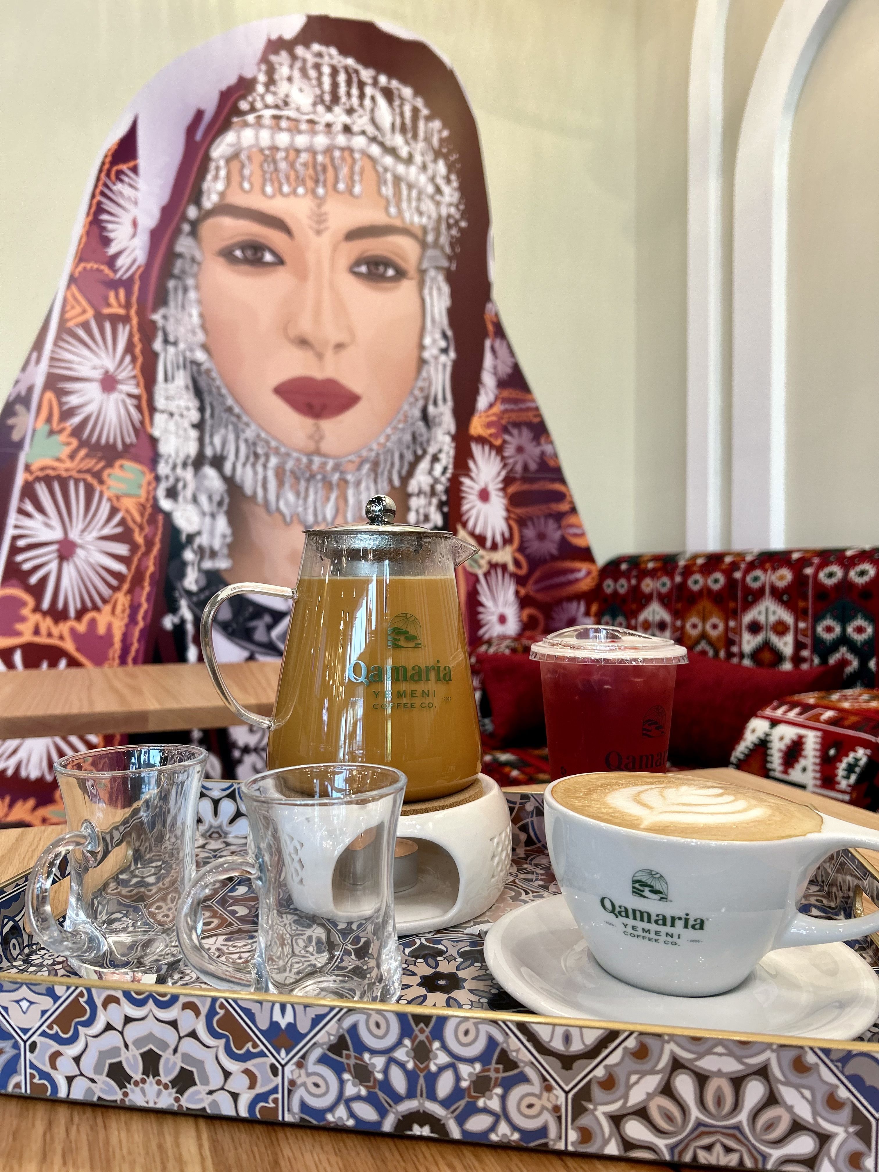Tray with Yemeni coffee set including a glass pot on a warmer, two glass cups, iced tea in a plastic cup, and a latte in a white cup on a table. Background features colorful mural of a woman in traditional attire.