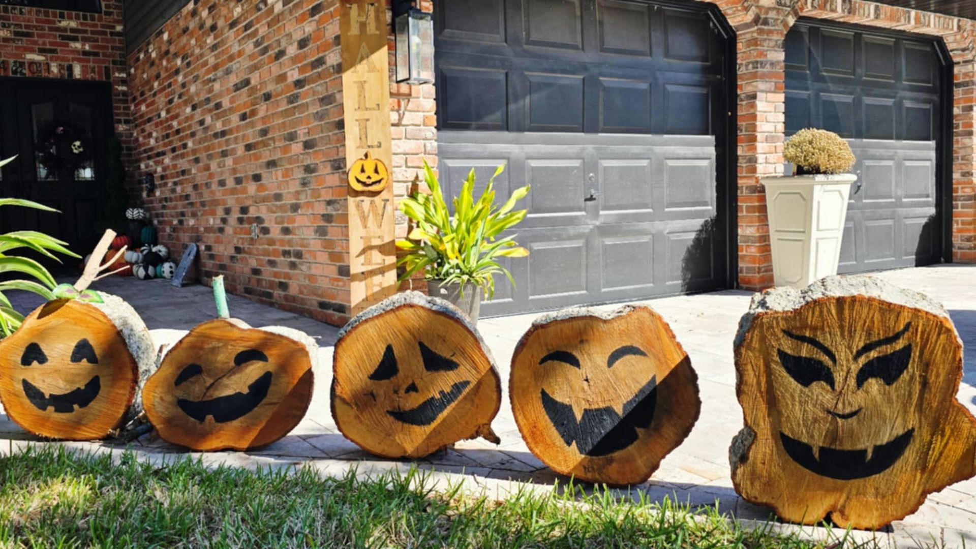 Five tree stumps lined up on a driveway, laying on their bark sides with the tree-ring sides facing the camera. Each stump has a Jack-o-lantern face drawn on.