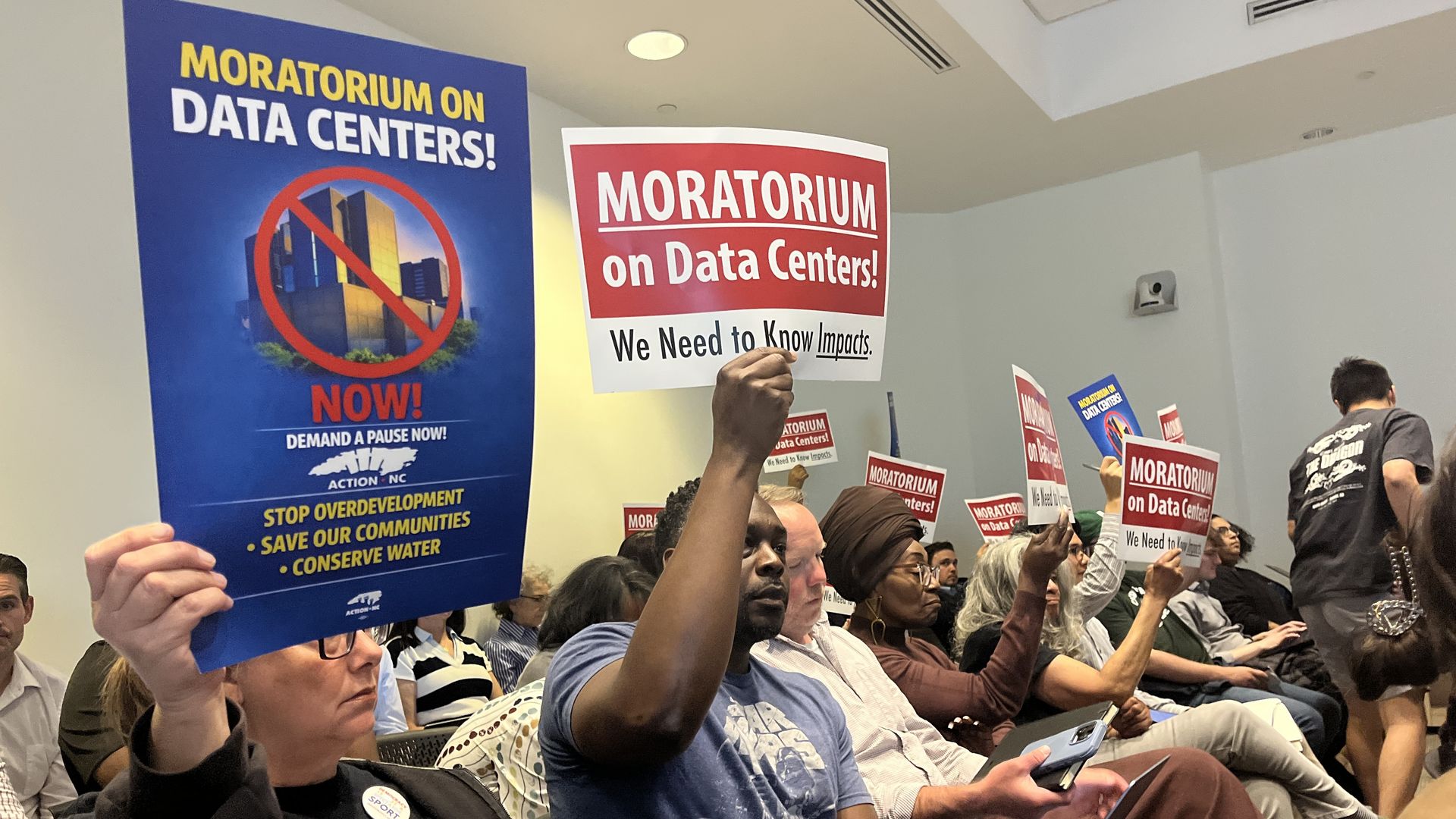 Crowd in a conference room holds protest signs: a large blue poster reading MORATORIUM ON DATA CENTERS! and a red sign reading MORATORIUM on Data Centers!; smaller signs say We Need to Know Impacts.