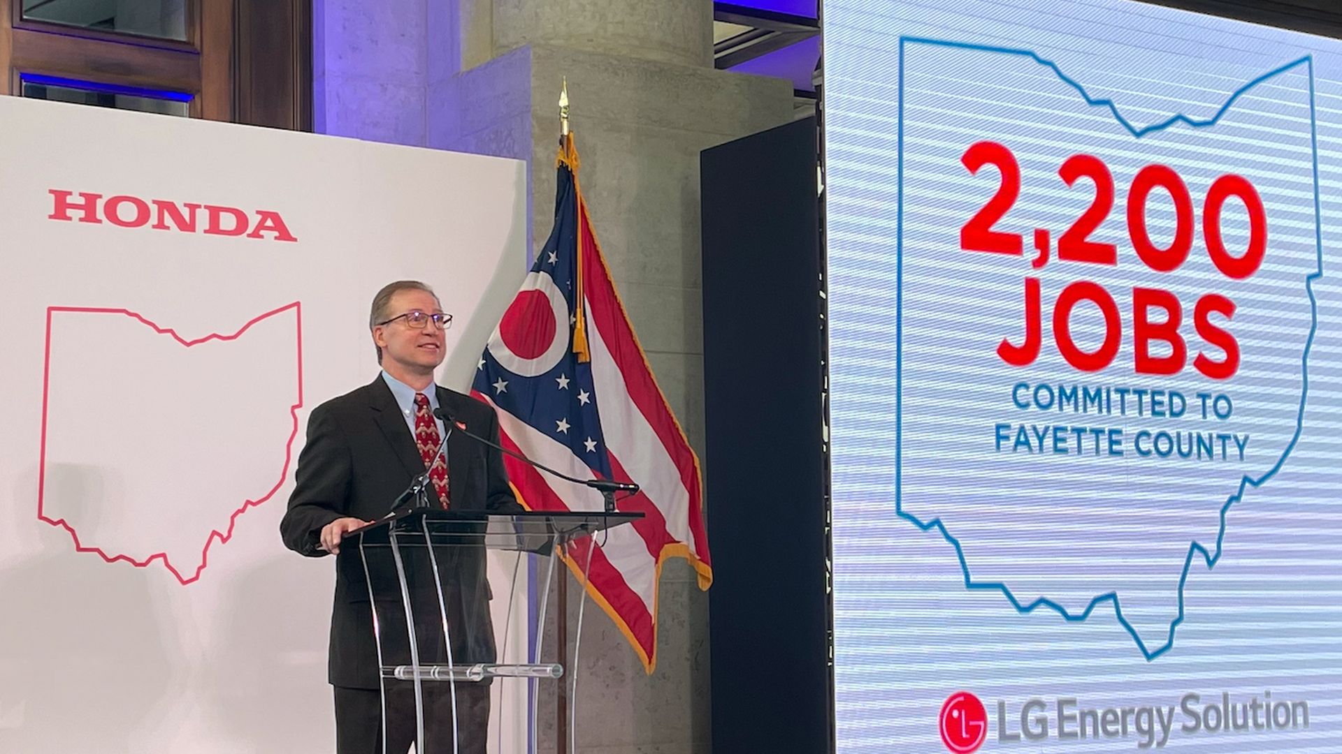 Bob Nelson, the executive vice-president of American Honda Motors, Co., Inc., addresses a crowd standing in front of an Ohio flag and Ohio Honda logo. 