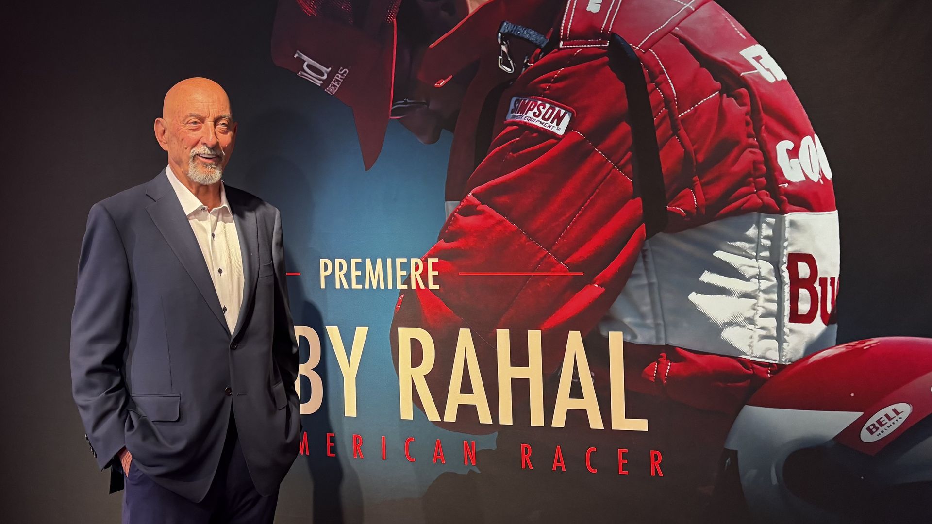 A bald man in a navy suit stands beside a large poster featuring a red racing suit with sponsor patches; the poster includes the words Premiere, By Rahal, and American Racer.