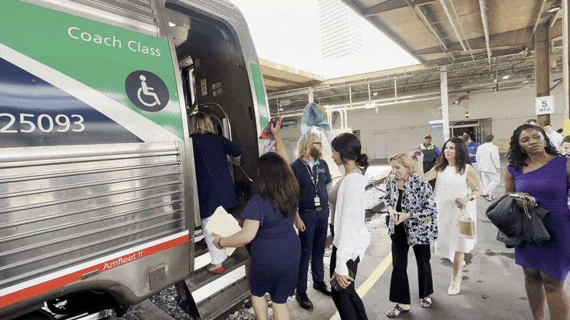 People boarding a silver and green coach class train at a station platform, some carrying bags and papers, with a 5 MPH sign and station ceiling visible above.