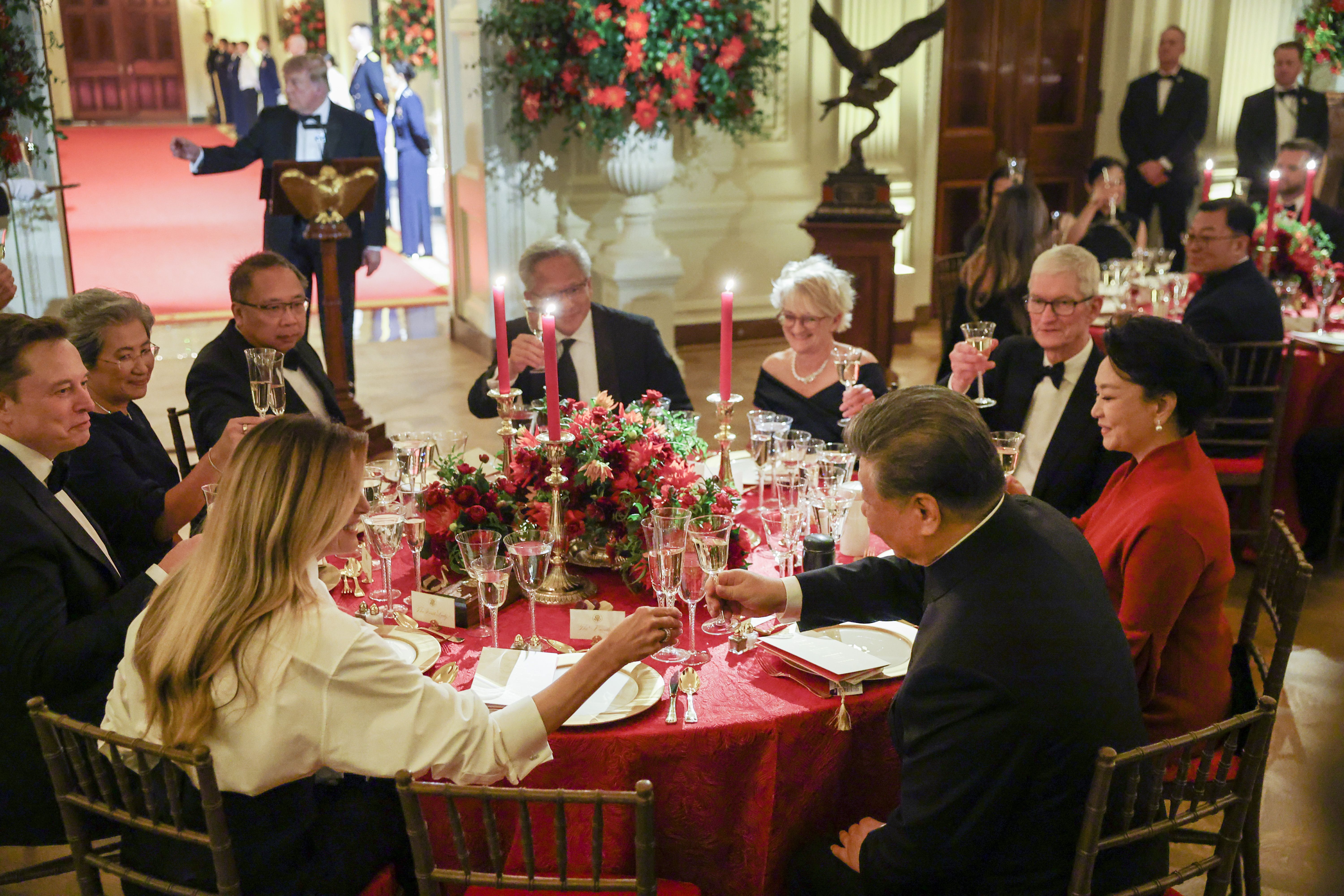 Guests in tuxedos and formal clothing — including first lady Melania Trump and President Xi Jinping — toast around a red tablecloth with pink candles and a lush floral centerpiece; a podium, behind which President Trump stands — and red carpet backdrop are visible.