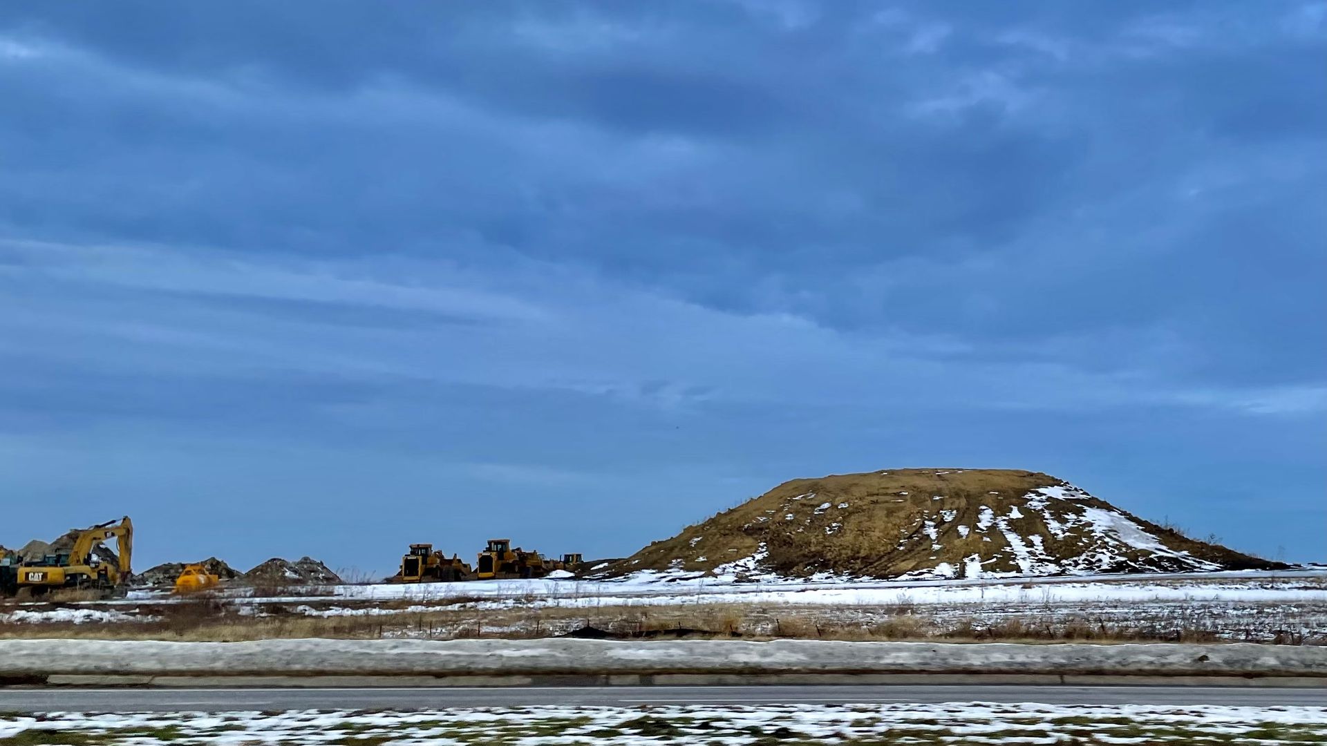 Construction site with yellow machinery and equipment near a large snow-dotted dirt mound under a cloudy blue sky in a wintery landscape.