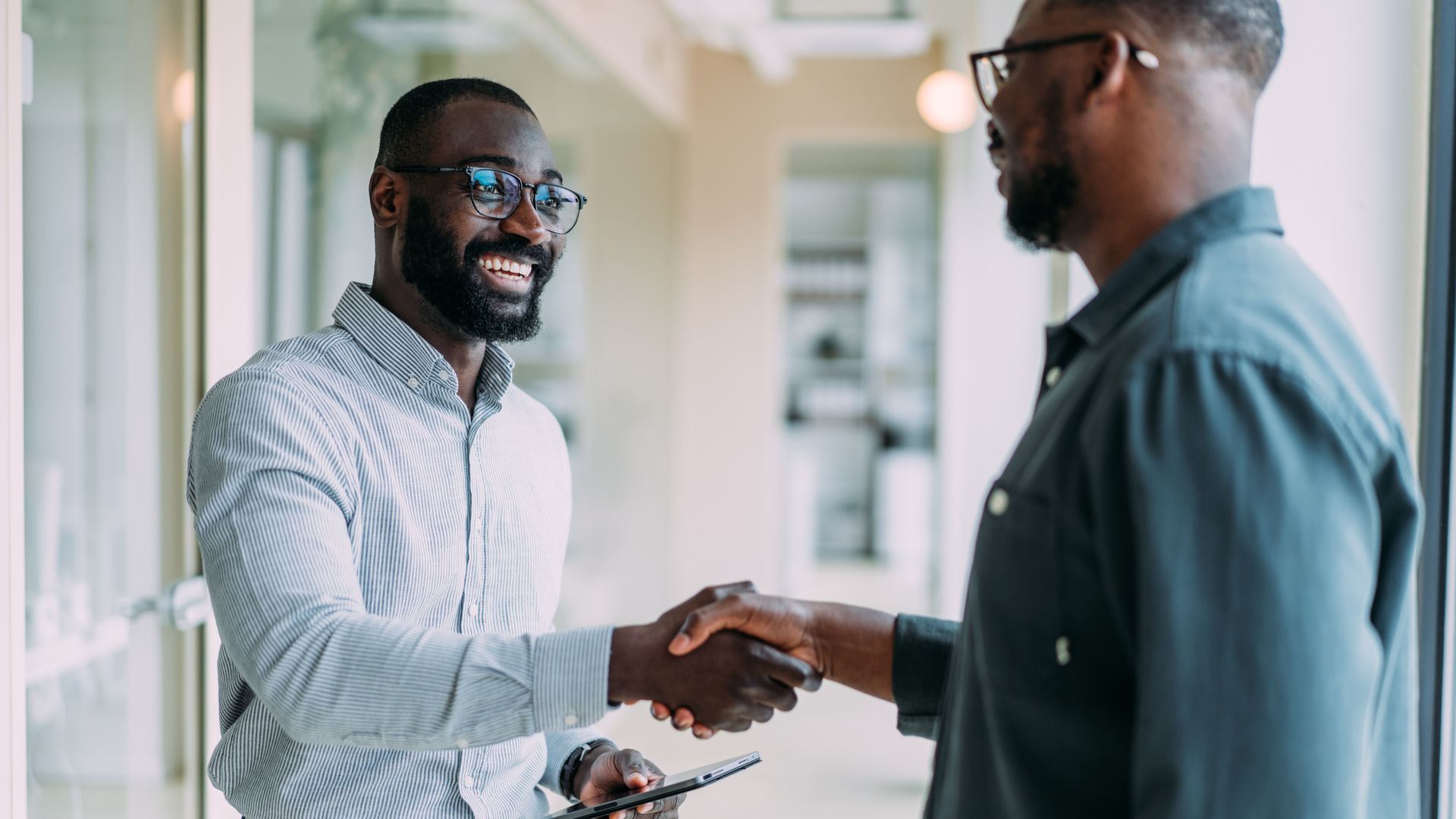 Two men, both wearing glasses, shake hands in a bright office. One holds a tablet and smiles, dressed in a striped shirt and dark pants; the other wears a dark shirt.