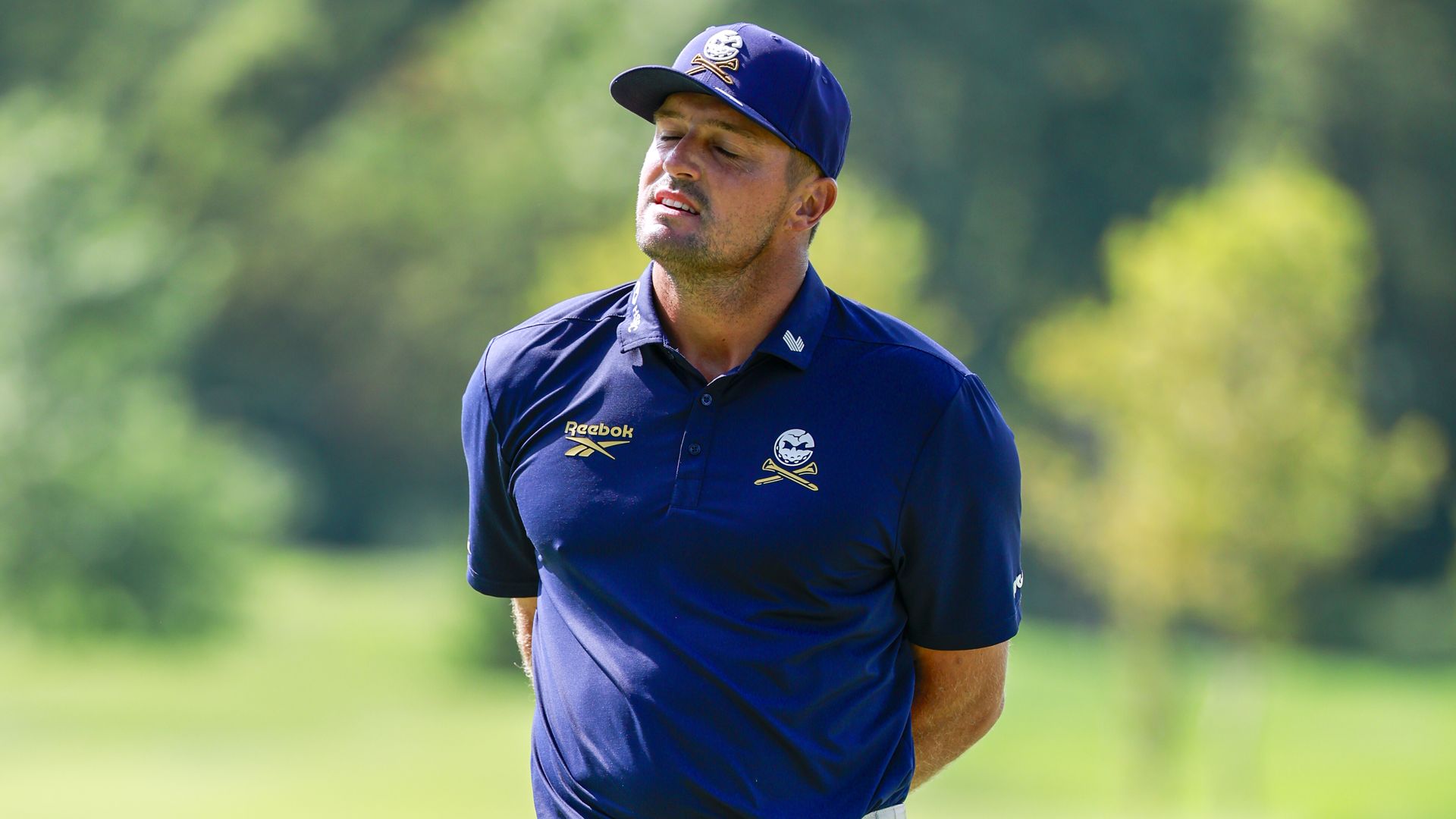 A golfer wearing a navy blue polo and matching cap stands outdoors on a sunny course, eyes closed, with a blurred green background.