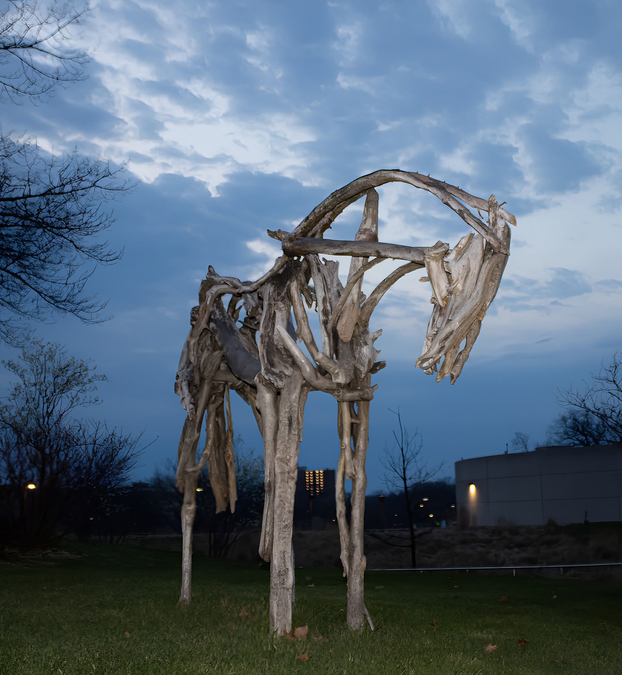 Silhouette of intertwined, pale wooden tree-skeleton sculpture forming circular arches in a grassy park, set against a blue sky with wispy clouds and leafless trees in the background.