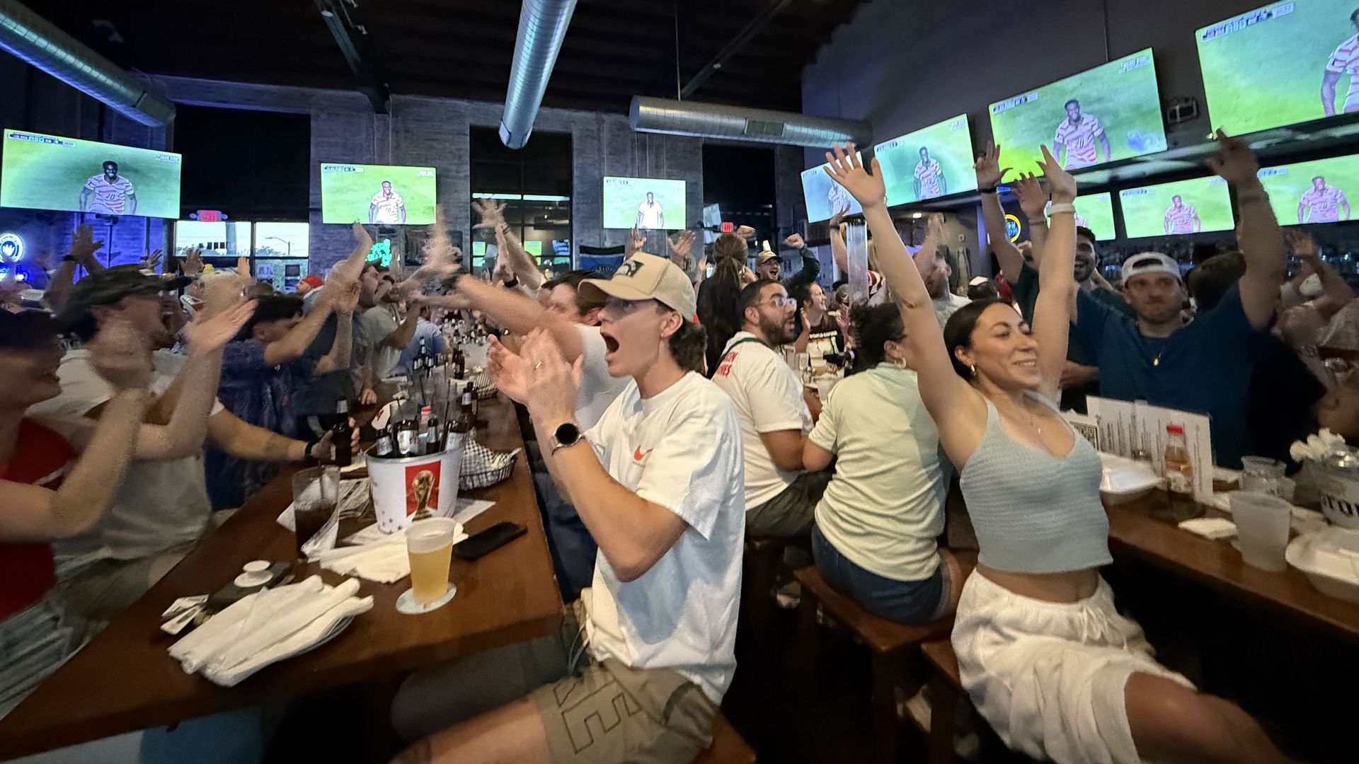 Busy indoor sports bar full of cheering people raising arms, long tables with drinks and plates, multiple green-screen TVs showing players, exposed ductwork, and warm string lights.