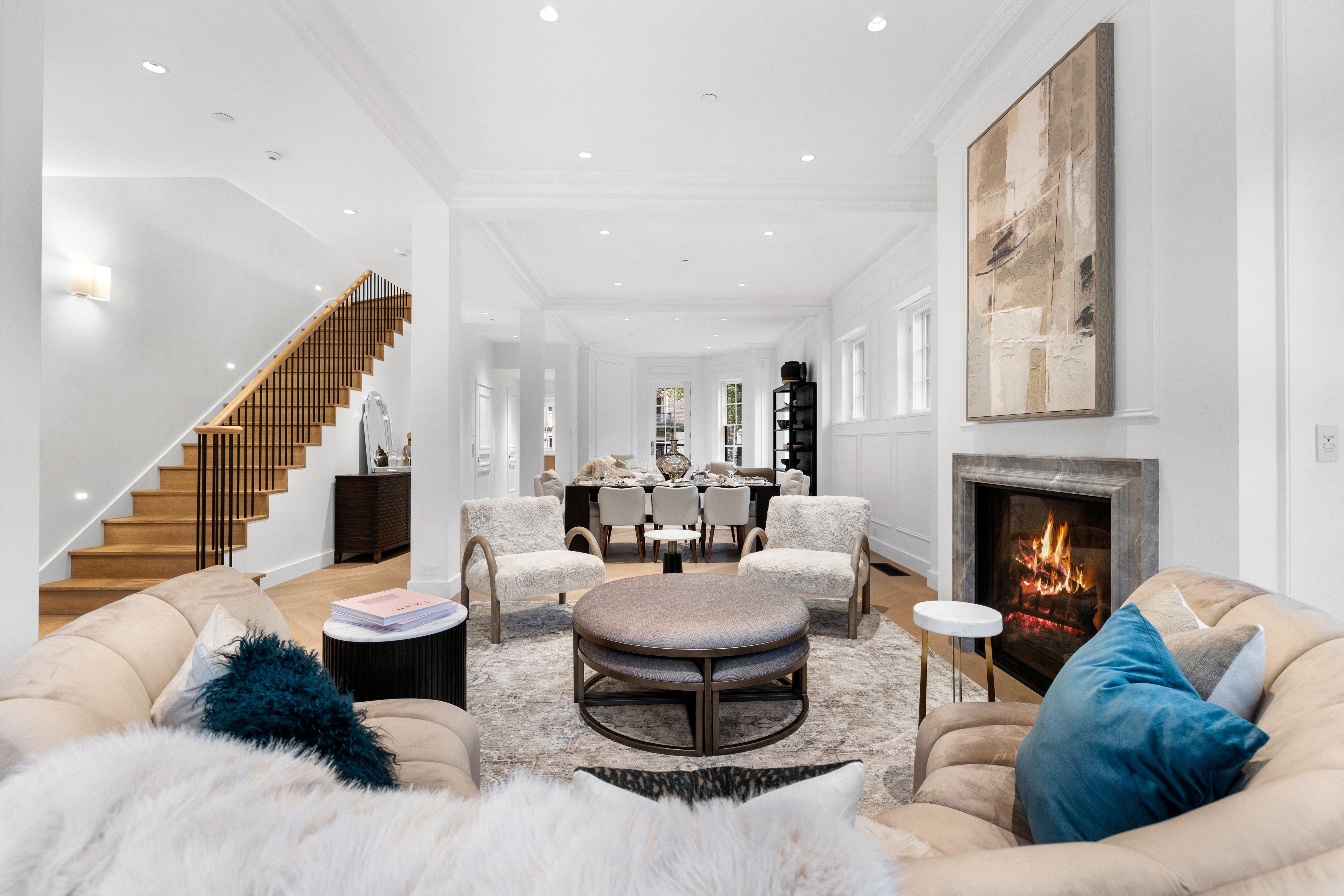 Bright modern living room with beige sofas, blue and white pillows, two furry chairs around a round ottoman, a lit fireplace beneath abstract art, wooden stairs, and a dining table in the back.