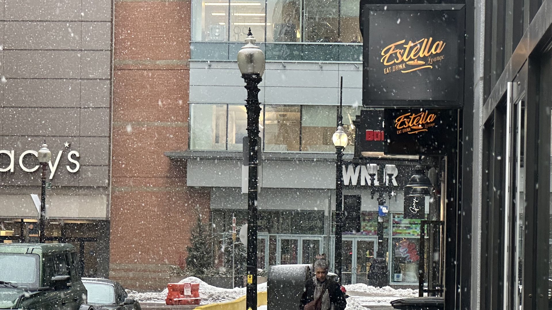 Snow falls on a city street where a person walks bundled in a scarf and coat near piled snow. Signs for Estella lounge and Macy's are visible along with parked cars and street lamps.
