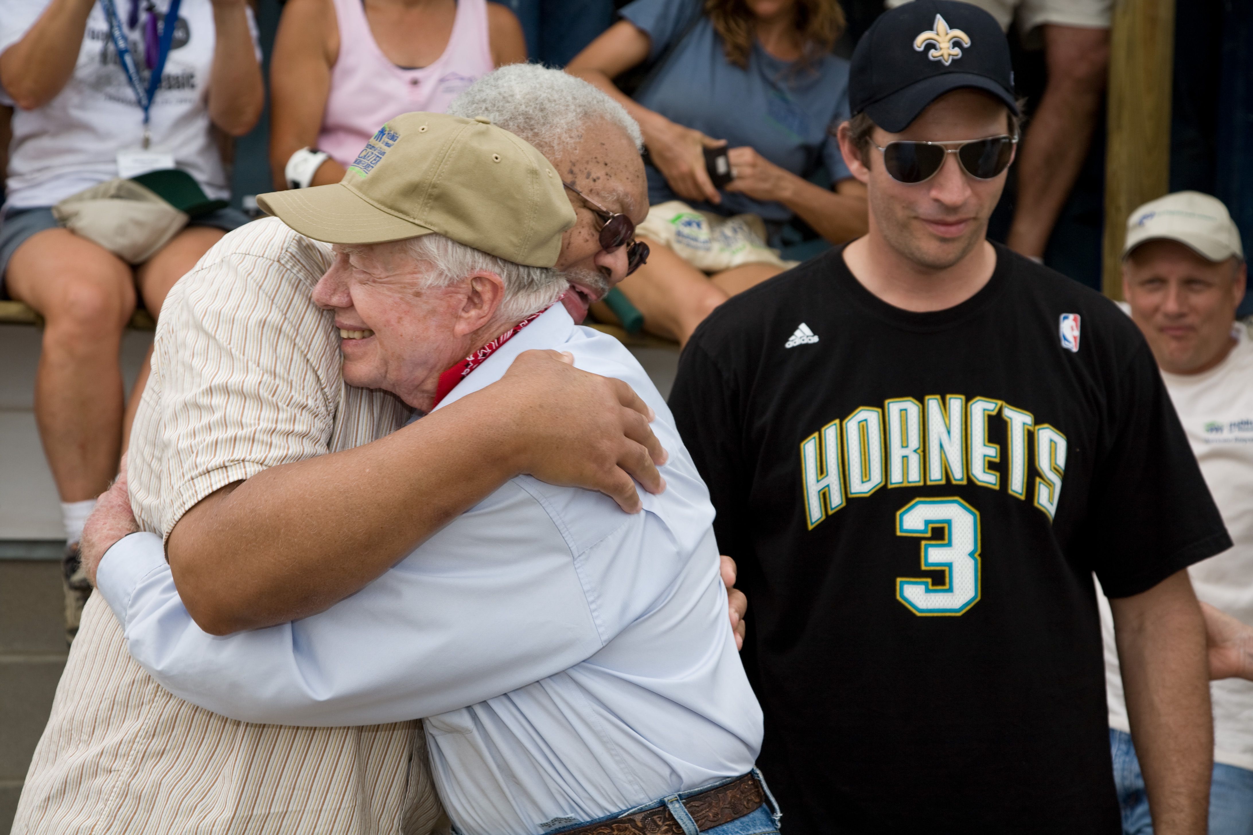 Jimmy Carter hugs a man in New Orleans, while another man in a Saints hat and a Hornets jersey stands nearby