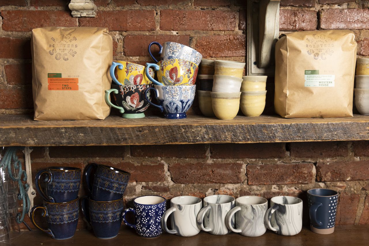Two brown coffee bags labeled Two Steps and Western House sit on a wooden shelf with colorful patterned mugs and yellow ceramic cups against a brick wall background.