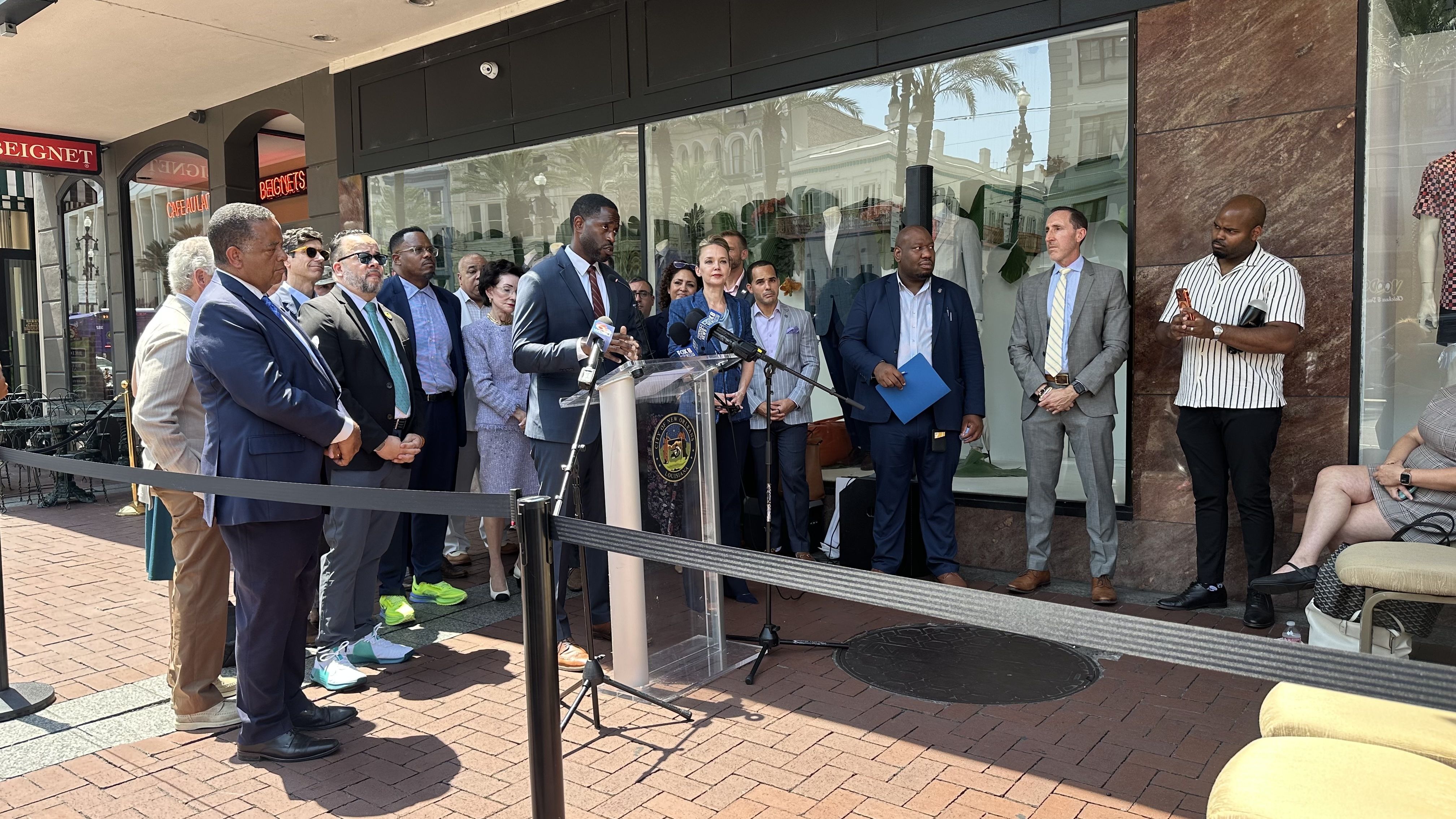 Group of officials and reporters outside a storefront press conference. A man in a dark suit speaks at a podium with the City seal; colleagues stand nearby as glass storefronts reflect palm trees and buildings.