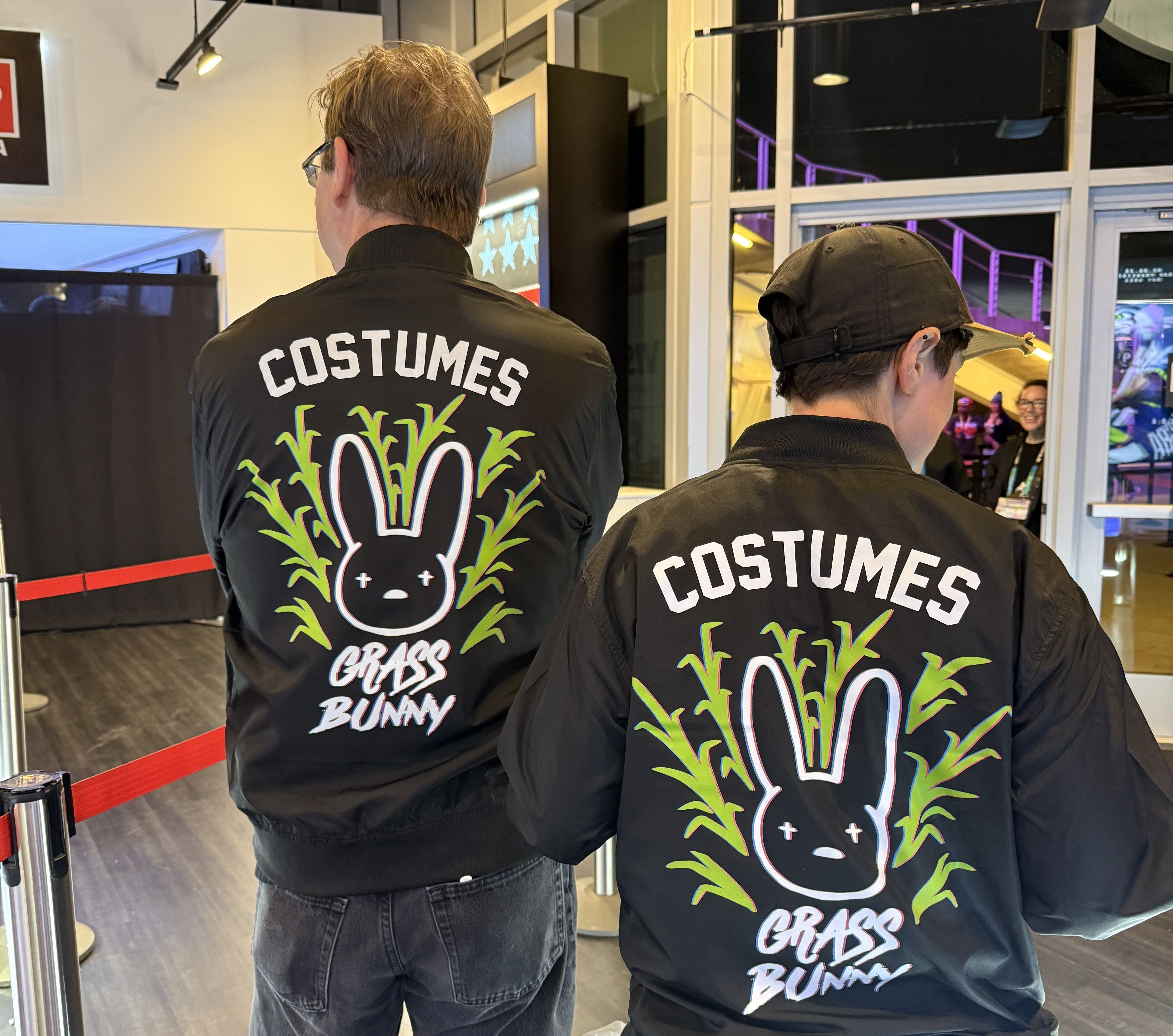 Two people wearing black jackets with a colorful design of a black bunny face surrounded by green grass and the words "COSTUMES" and "GRASS BUNNY" on the back.