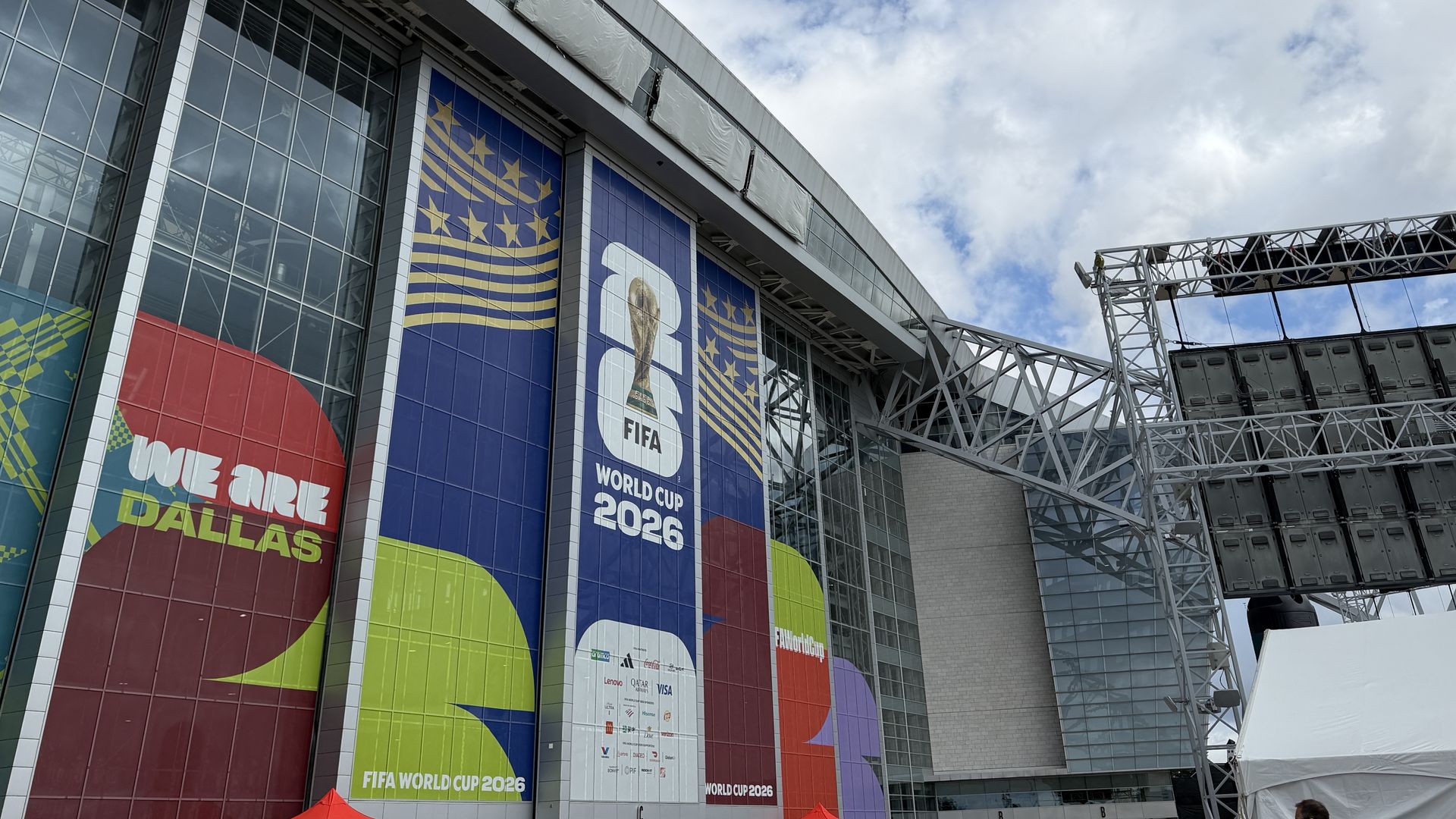 Modern stadium facade with large color banners: "FIFA World Cup 2026" and "WE ARE DALLAS". Blue panels with star patterns, glass walls, and a metal truss framework against a cloudy sky.