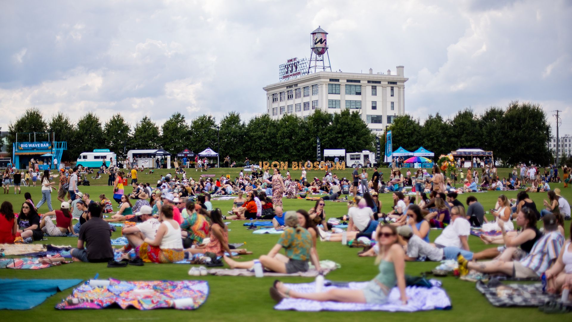A crowded city park festival: people sit on colorful blankets on the grass, with a white building and water tower in the distance, framed by trees and tents.
