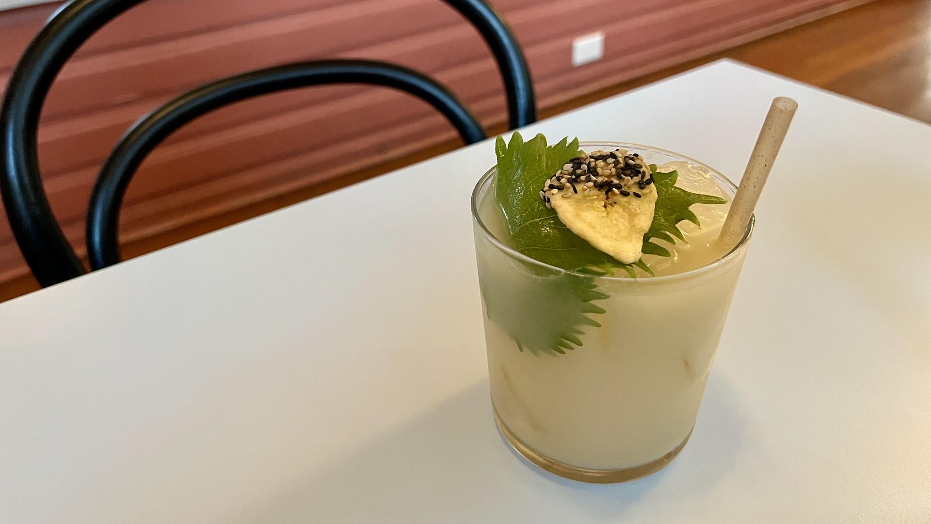 A yellow-colored cocktail is garnished with pineapple, greenery and sesame seeds and is seen on a white table with a pink wall in the background.