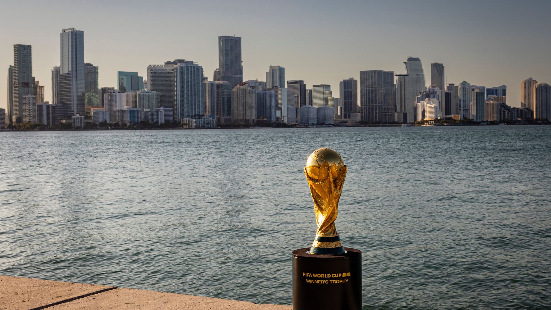 Golden FIFA World Cup winner's trophy on a black pedestal by waterfront with a city skyline and calm water under a clear sky in the background.