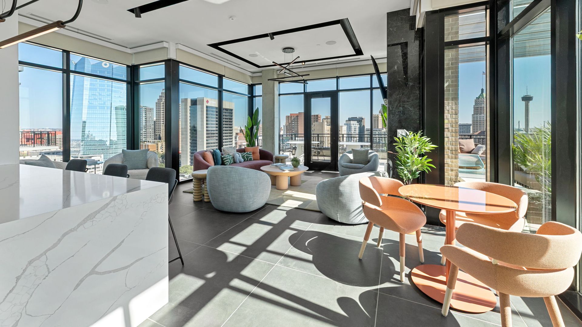 A bright, modern living room with large floor-to-ceiling windows showing the San Antonio skyline and the Tower of the Americas. There are gray and coral chairs, a pink couch, a round wooden coffee table, and some plants at The Continental Residences.
