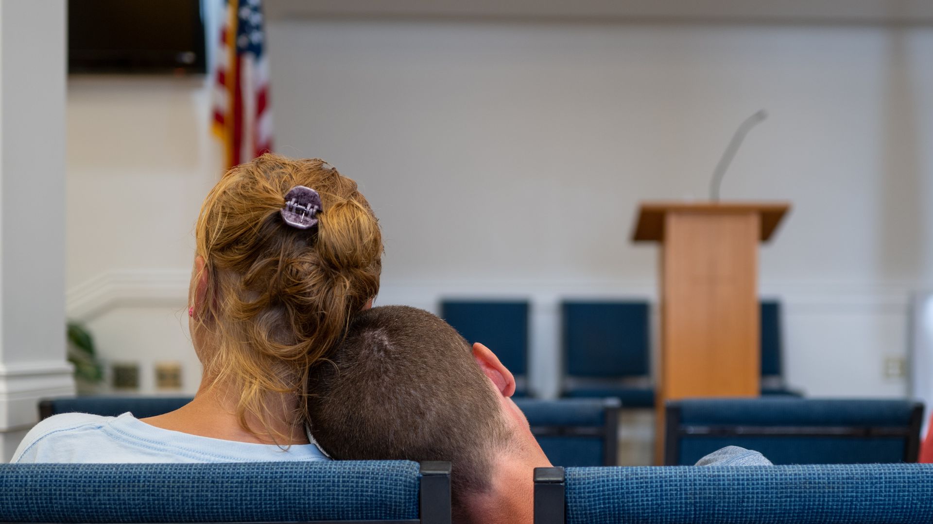 Two people sitting on blue chairs in a room with an American flag, a podium with a microphone, and empty chairs in the background. One person's head rests on the other's shoulder.