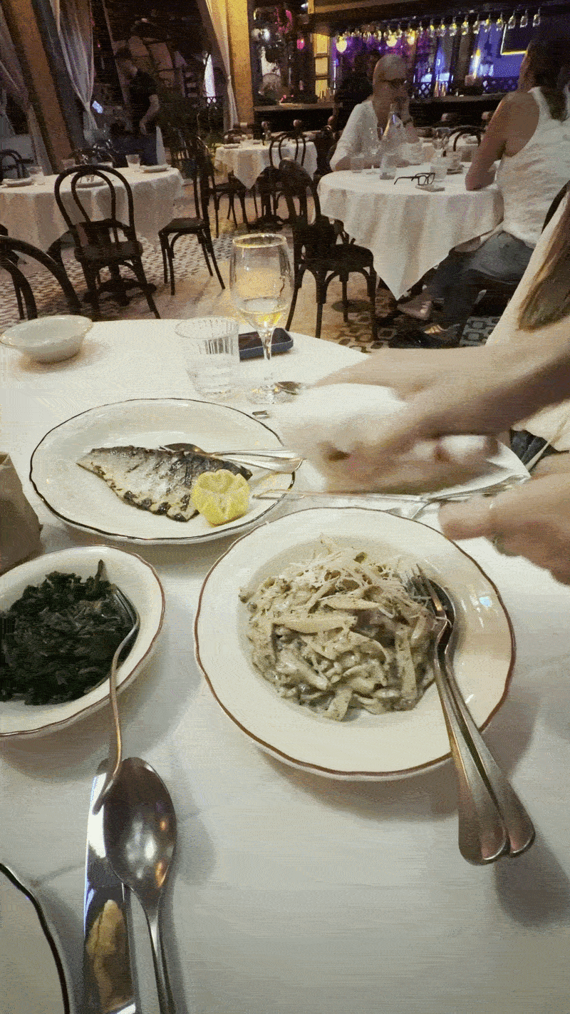 Restaurant table with white cloths; plates of grilled fish with lemon, creamy pasta, and greens. A glass of white wine; blurred diners in the background as someone serves pasta.