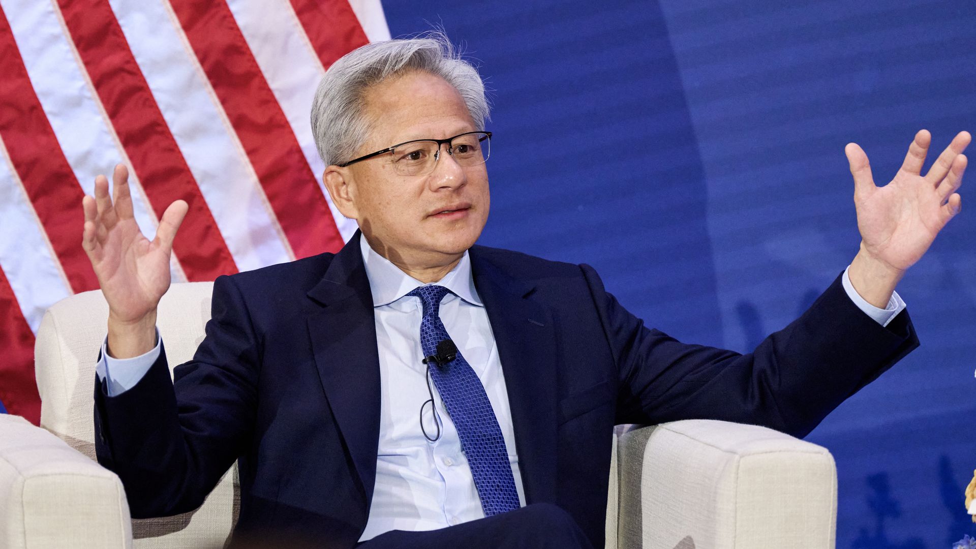 A person with gray hair and glasses, wearing a dark suit and blue tie, sits and gestures with both hands raised. An American flag and blue backdrop are visible behind him.