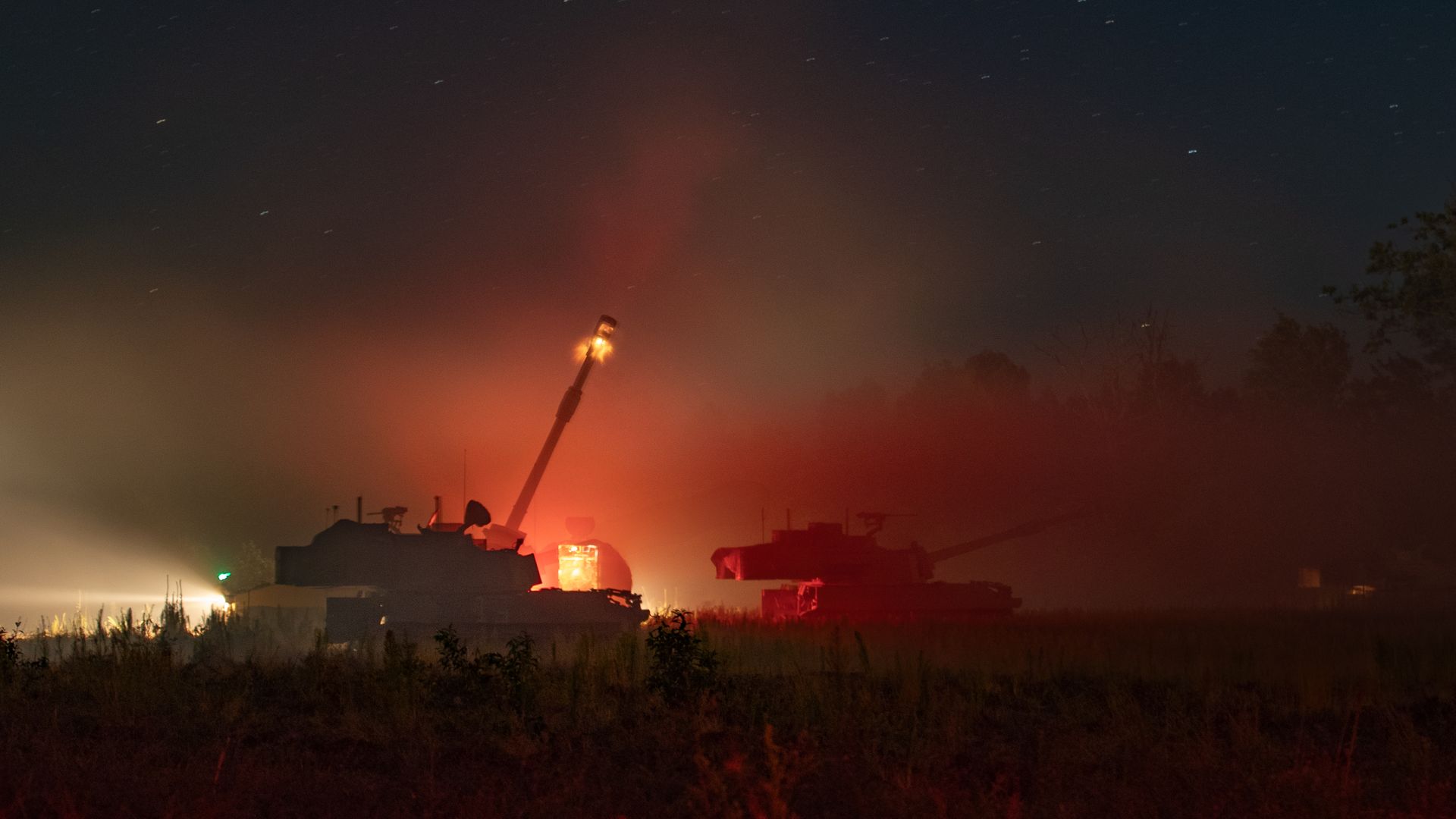 Self-propelled howitzers are seen at night in a field. One if shooting, with a fireball exiting the barrel.