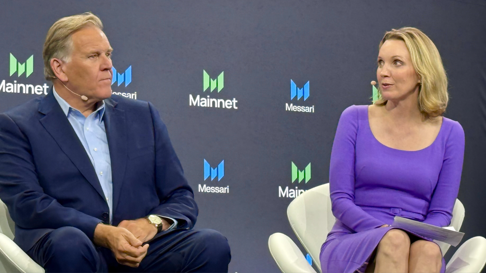 A man and a woman talking on stage at a conference. He's in a blue suit, she's in a purple dress.