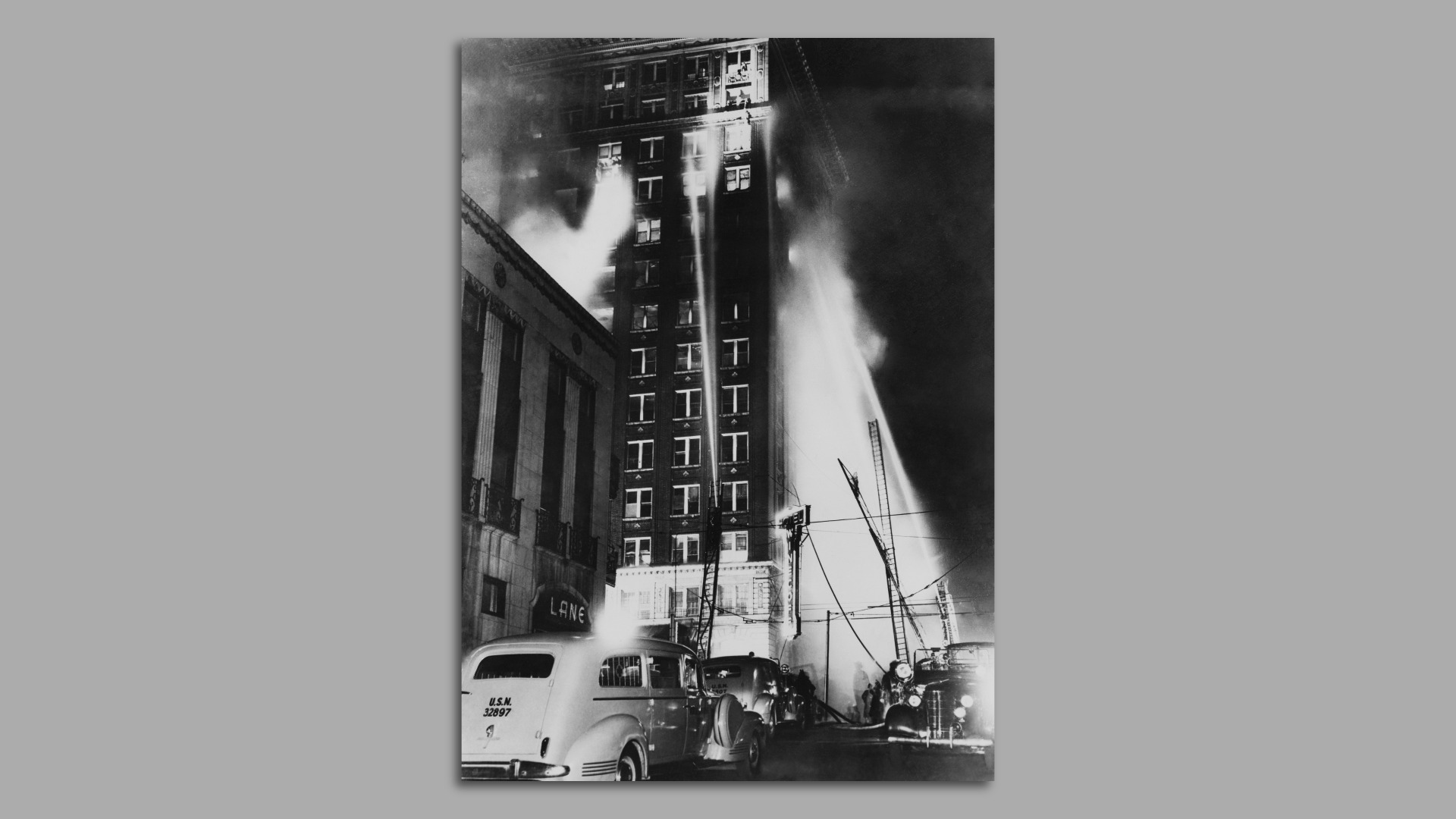 A black and white photo of firefighters spraying water to the flaming upper floors of a 15-story hotel