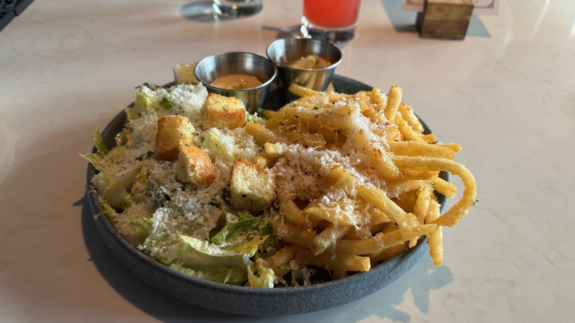 Dark bowl filled with crispy French fries and romaine salad sprinkled with parmesan; two small metal cups of dipping sauce sit nearby on a light table.