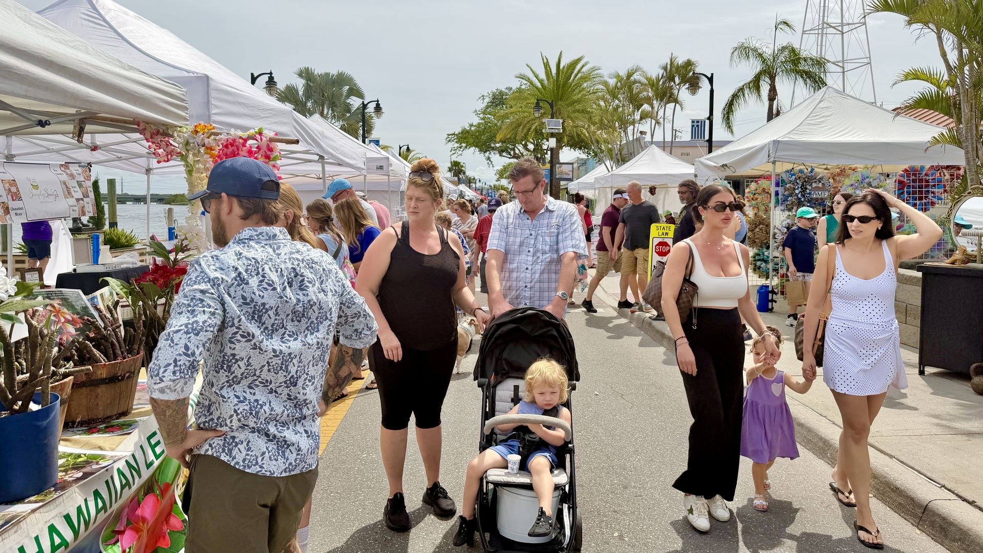 Busy outdoor market with white vendor tents along a sunny street, tropical plants and hibiscus leis, people strolling, a woman pushing a stroller with a child, and a tall metal tower in the background.