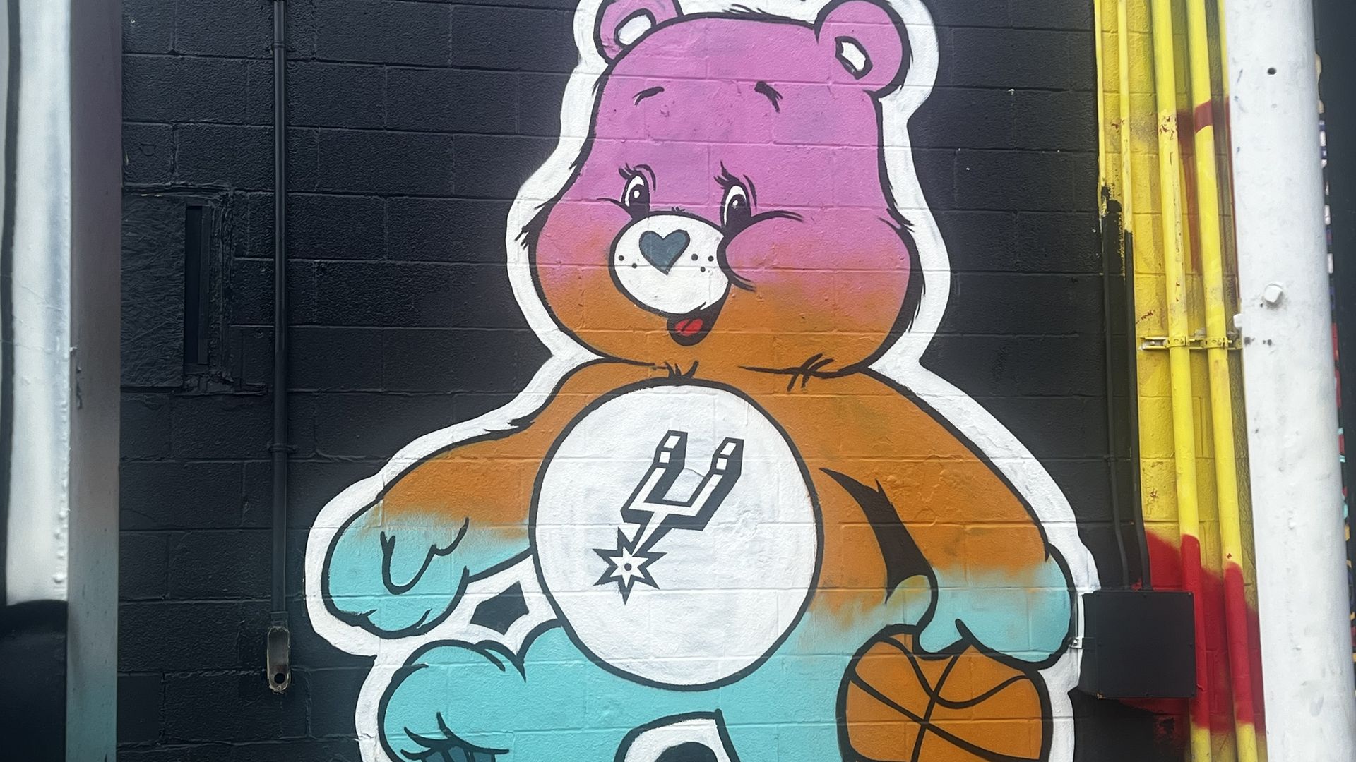 Colorful graffiti mural of a large pink cartoon bear with a blue heart-shaped nose, white outline, orange belly and teal paw, holding a basketball. Black wall backdrop with yellow pipes on the right.