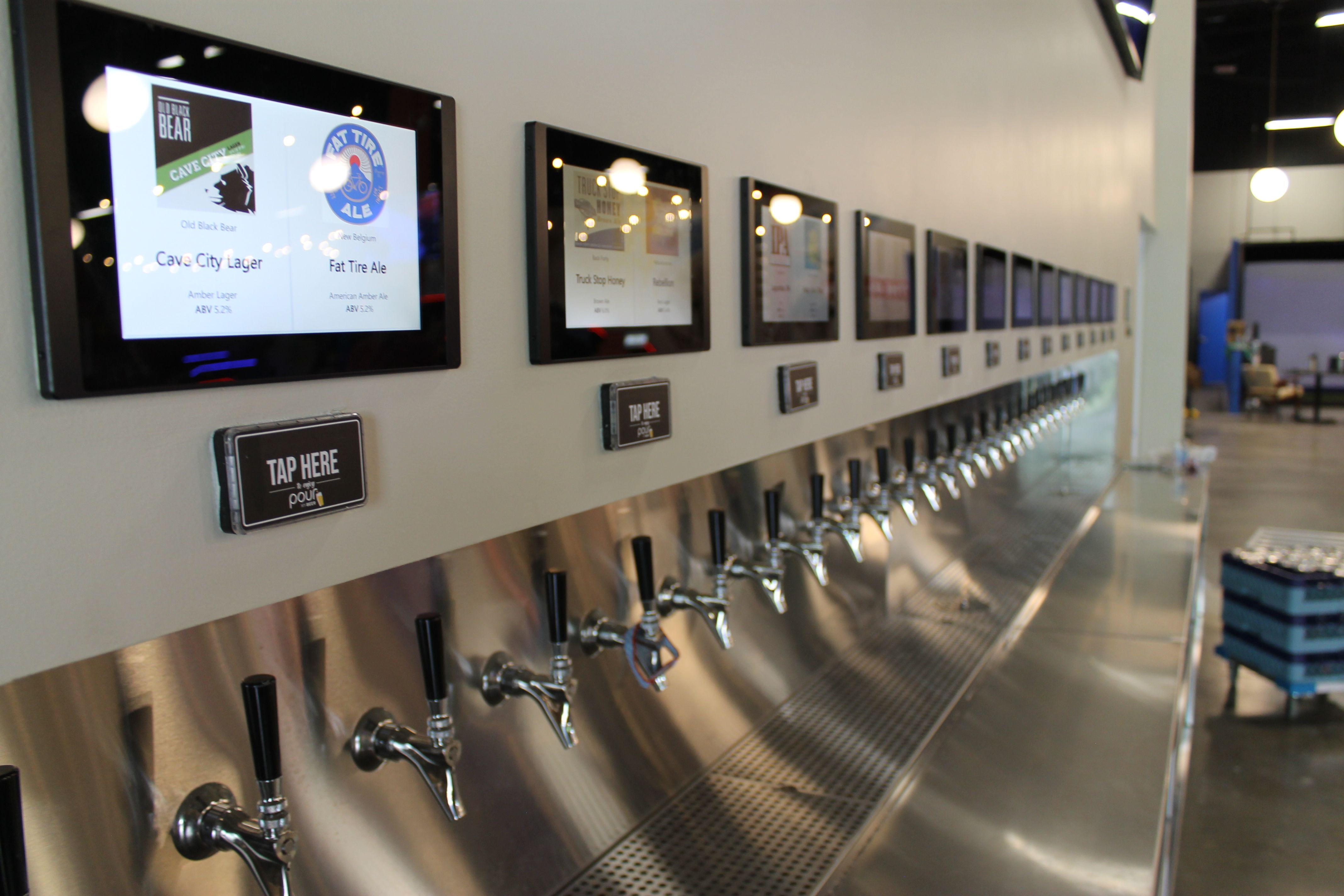 A long row of beer taps with digital screens above each one displaying beer names and logos like Cave City Lager and Fat Tire Ale in a modern bar setting.