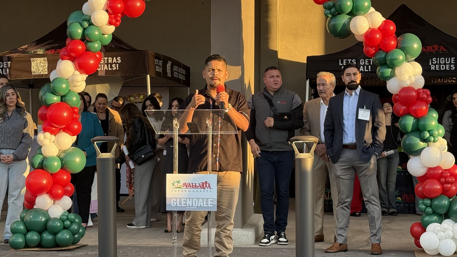 An outdoor event with a man at a podium speaking into a red microphone, flanked by red, white, and green balloon arches. People stand behind him near black tents; a Glendale sign is visible.