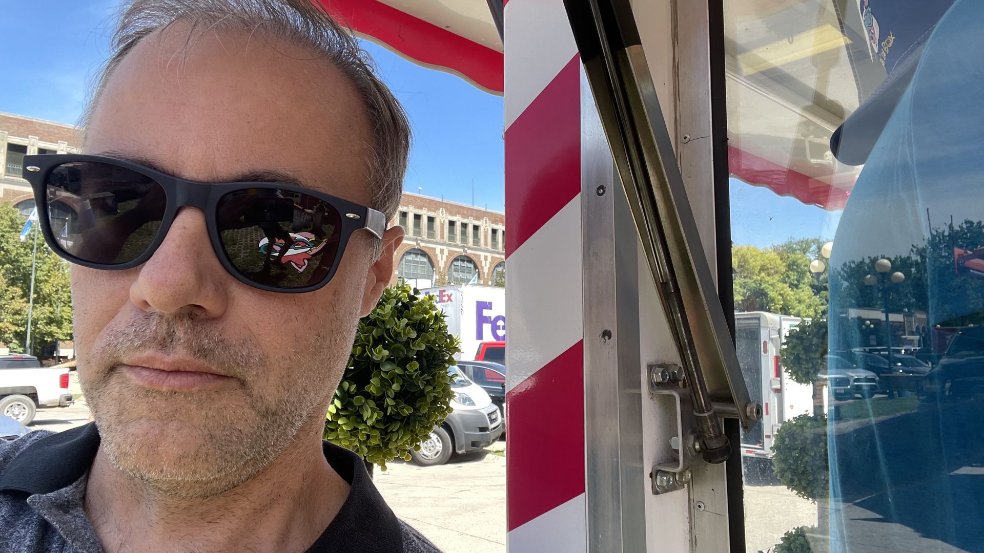 Man wearing black sunglasses and a dark shirt standing outside near a red and white striped pole, with parked cars and a brick building under a blue sky in the background.